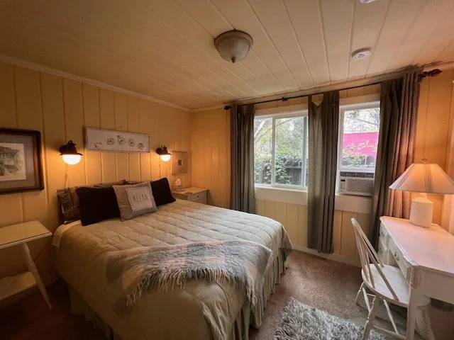 A cozy bedroom with a bed, nightstands, and lamps, featuring beige walls, a small window with curtains, and a window air conditioning unit.