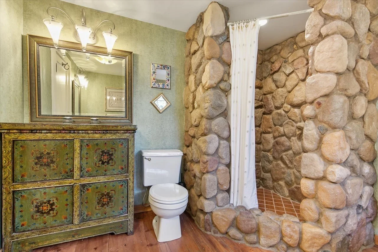 A rustic bathroom featuring a vintage green chest of drawers, a mirror, a toilet, and a stone shower enclosure with a white curtain, wood and stone accents, and a green wall.