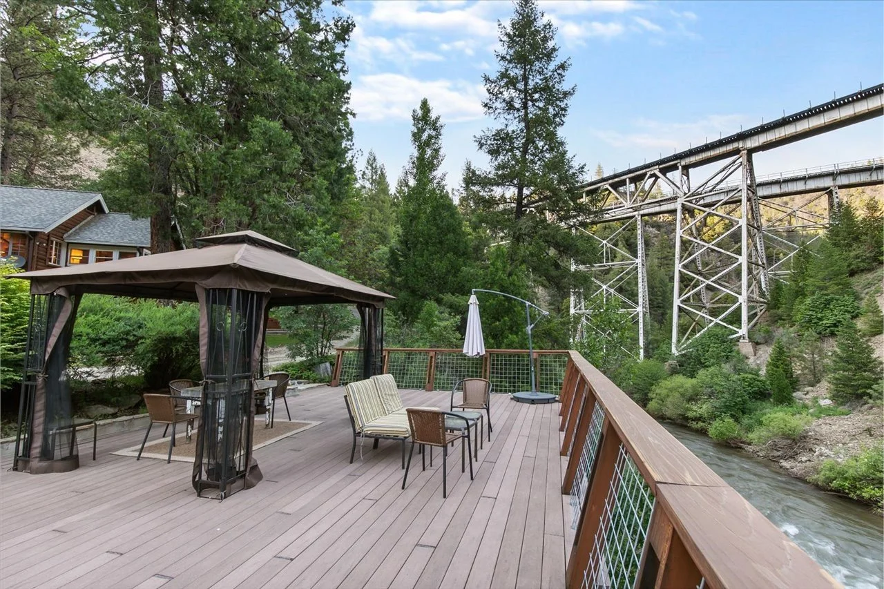 Outdoor wooden deck with chairs, tables, and a patio umbrella next to a river, overlooking a forested area and a large steel bridge.