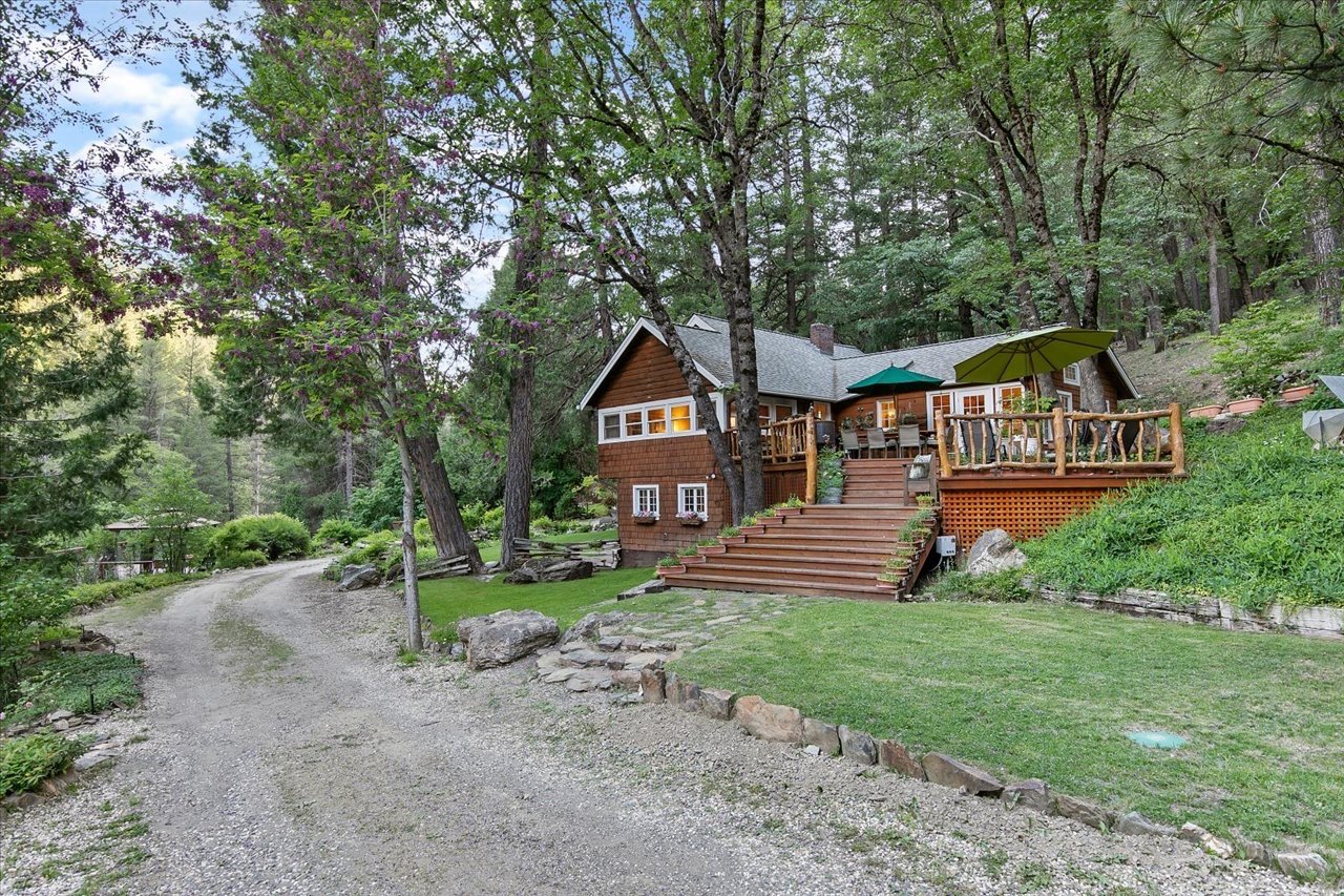 A rustic house with wooden steps leading up to a deck, surrounded by green trees and a gravel driveway.