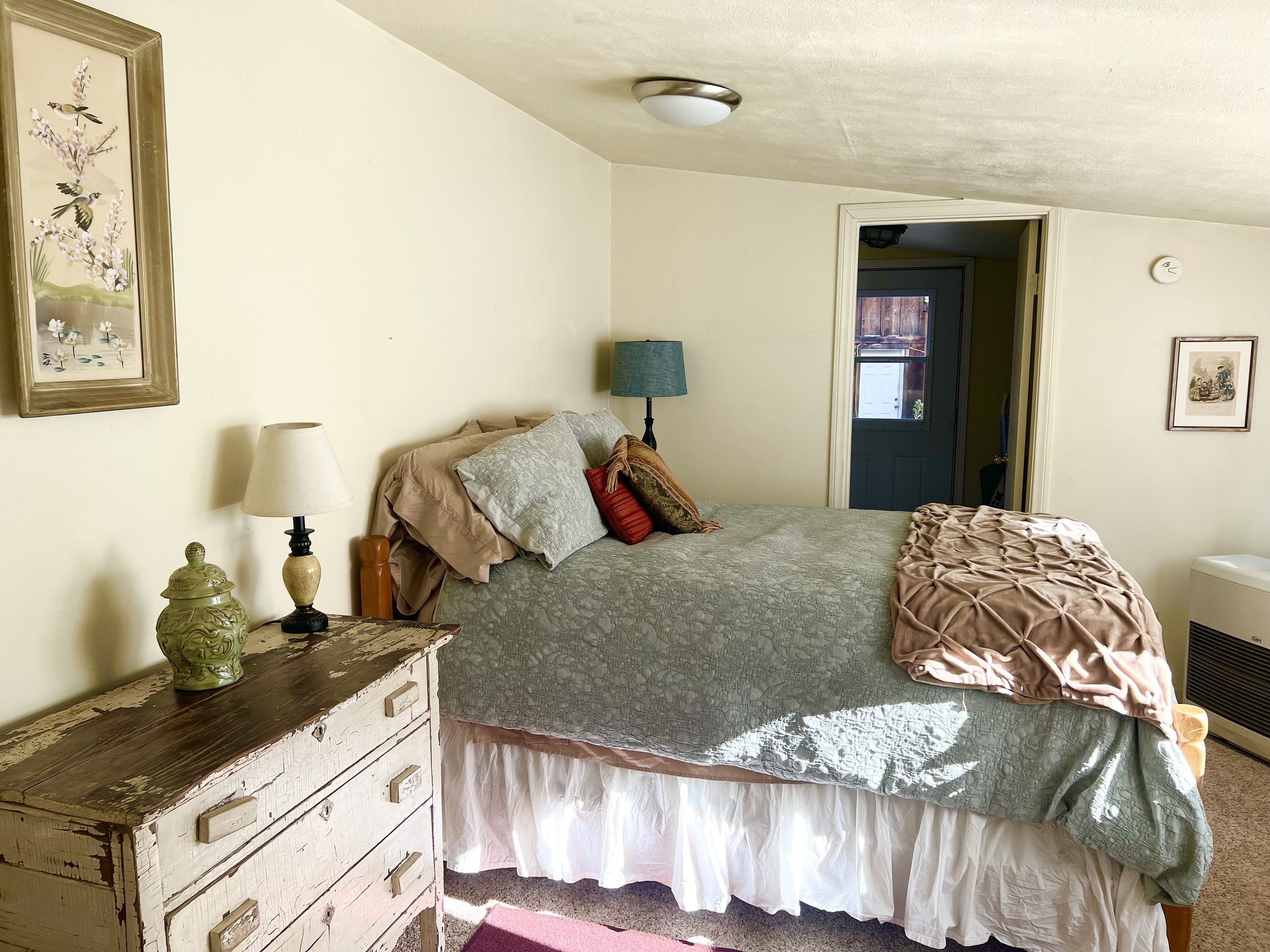 A bedroom with a wooden dresser, bed with patterned bedding, pillows, and a blanket, two lamps, wall art, and a door leading to an outside area.