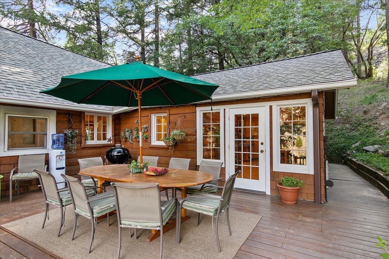Outdoor patio area with wooden deck, round table, several chairs, a green patio umbrella, potted plants, a water dispenser, and a grill set against a wooden house with multiple windows, surrounded by trees.