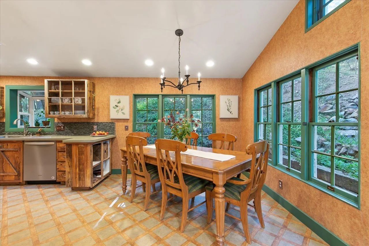 A cozy dining area with a wooden table and six matching chairs, a floral arrangement in the center, green framed windows showing a view of rocks and greenery outside, and a corner kitchen with wooden cabinets and a refrigerator.