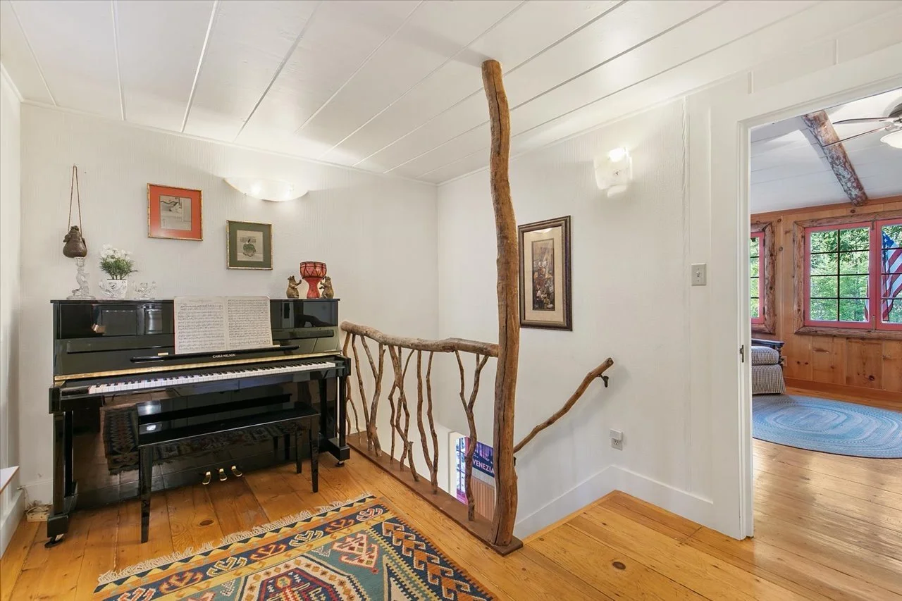 Interior view of a hallway with a black upright piano decorated with a music sheet, small statues, and a red lamp. Wooden stick railing runs along a staircase with a natural wood handrail and a twisted wooden post. The floor is hardwood with a printe