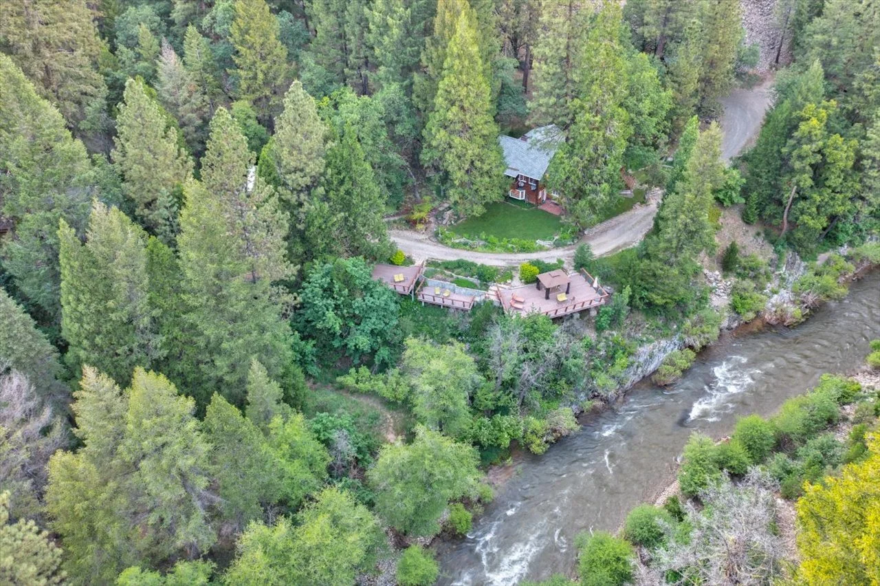 Aerial view of a house and multiple decks surrounded by dense green forest, with a river flowing along the edge of the property.