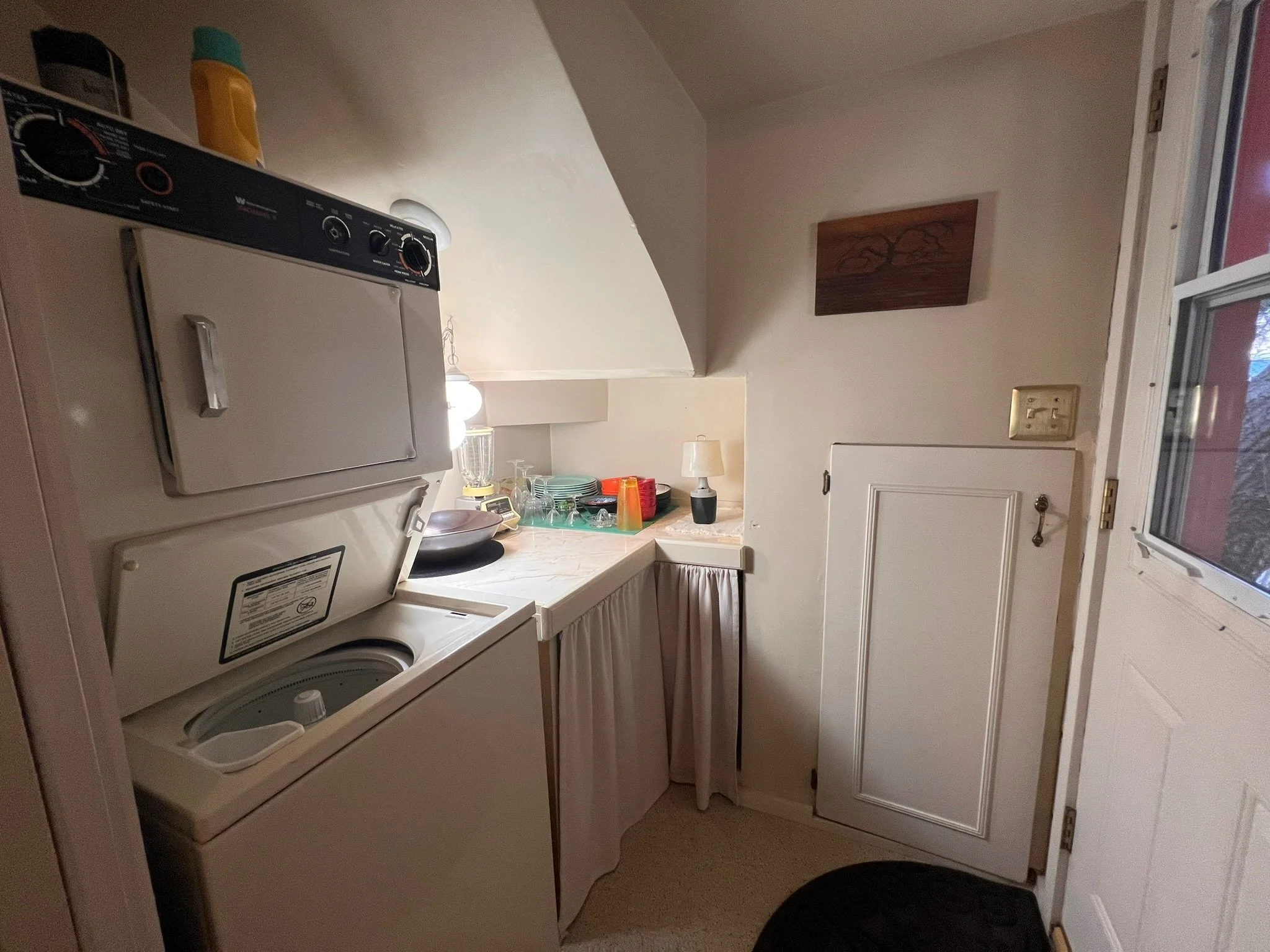 A laundry area with a stacked washing machine and dryer, a small countertop with dishes and glasses, a small lamp, and wall decorations. There is a door on the right side.