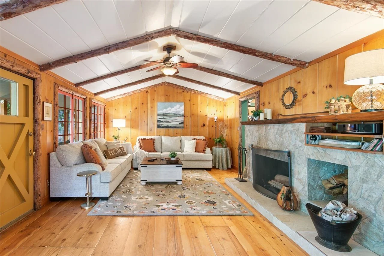 Cozy living room with wood-paneled walls, a stone fireplace, and a white sofa with orange and beige pillows. There are windows with red frames, a colorful area rug, and a ceiling fan with wooden blades.