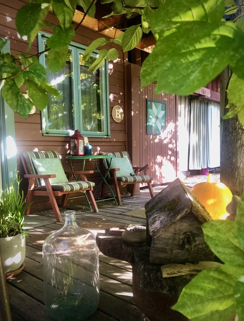 Cozy outdoor patio with wooden deck, two striped chairs with green cushions, a small green table, a lantern, a potted plant, a large glass jar, a pumpkin, and logs, shaded by leafy trees with the house's brown wooden siding and windows in the background.