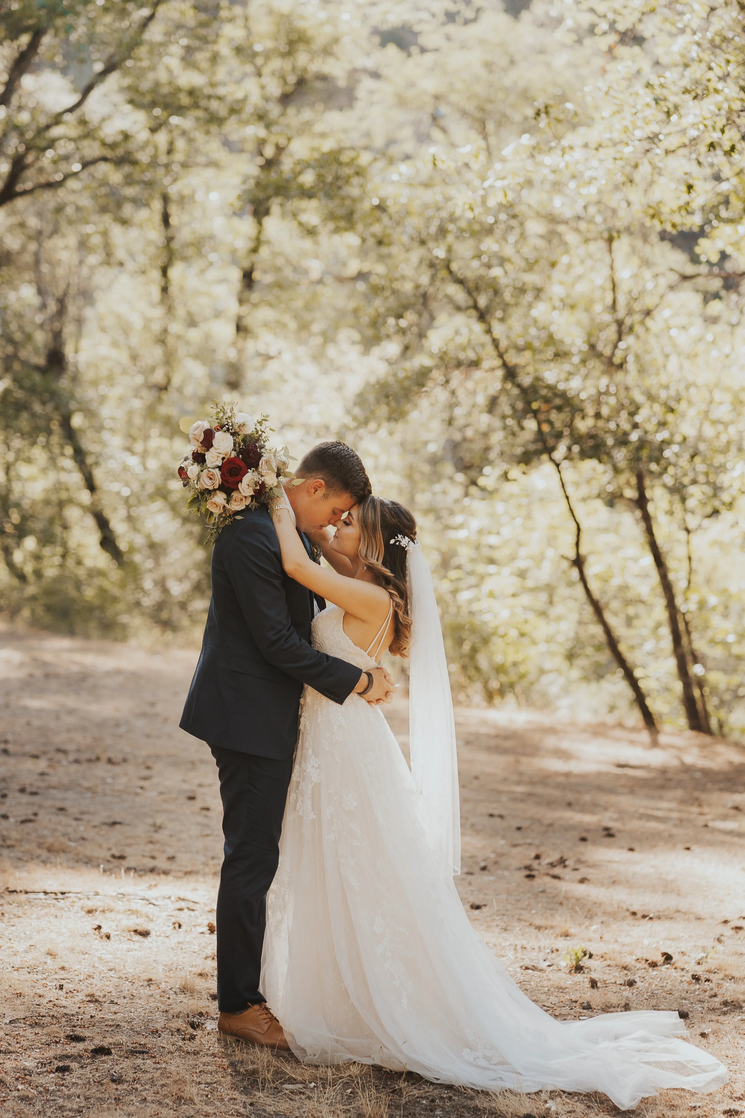 A bride and groom embrace outdoors on their wedding day, surrounded by trees with sunlight filtering through.
