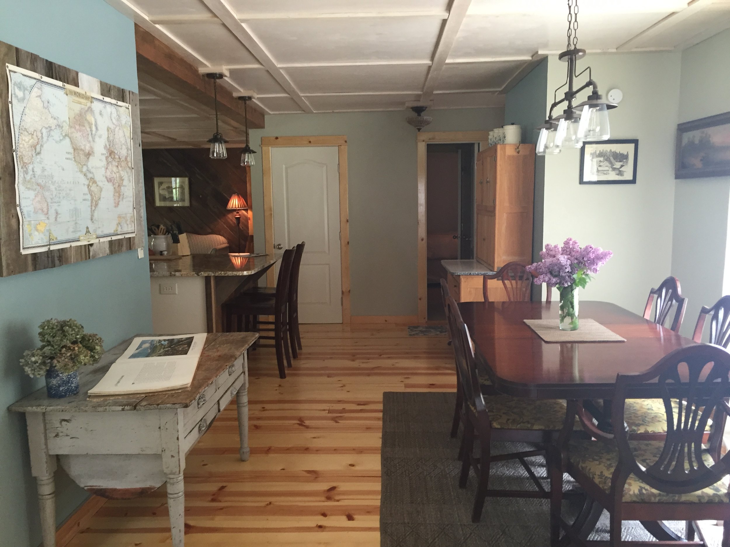 A dining room with a dark wood table and six matching chairs, a vase of purple flowers on the table, a small white side table with a potted plant and open book, a wooden cabinet, a ceiling light fixture, and pictures on the walls.