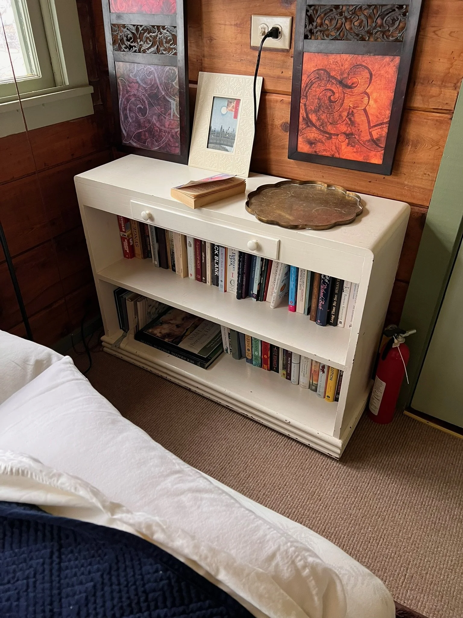 White bookshelf with two shelves filled with books, a brass tray, a framed picture, a stack of books, a decorative white frame, and a brass tray on top. Behind the bookshelf are two framed decorative art pieces and a wooden wall, with an electrical o