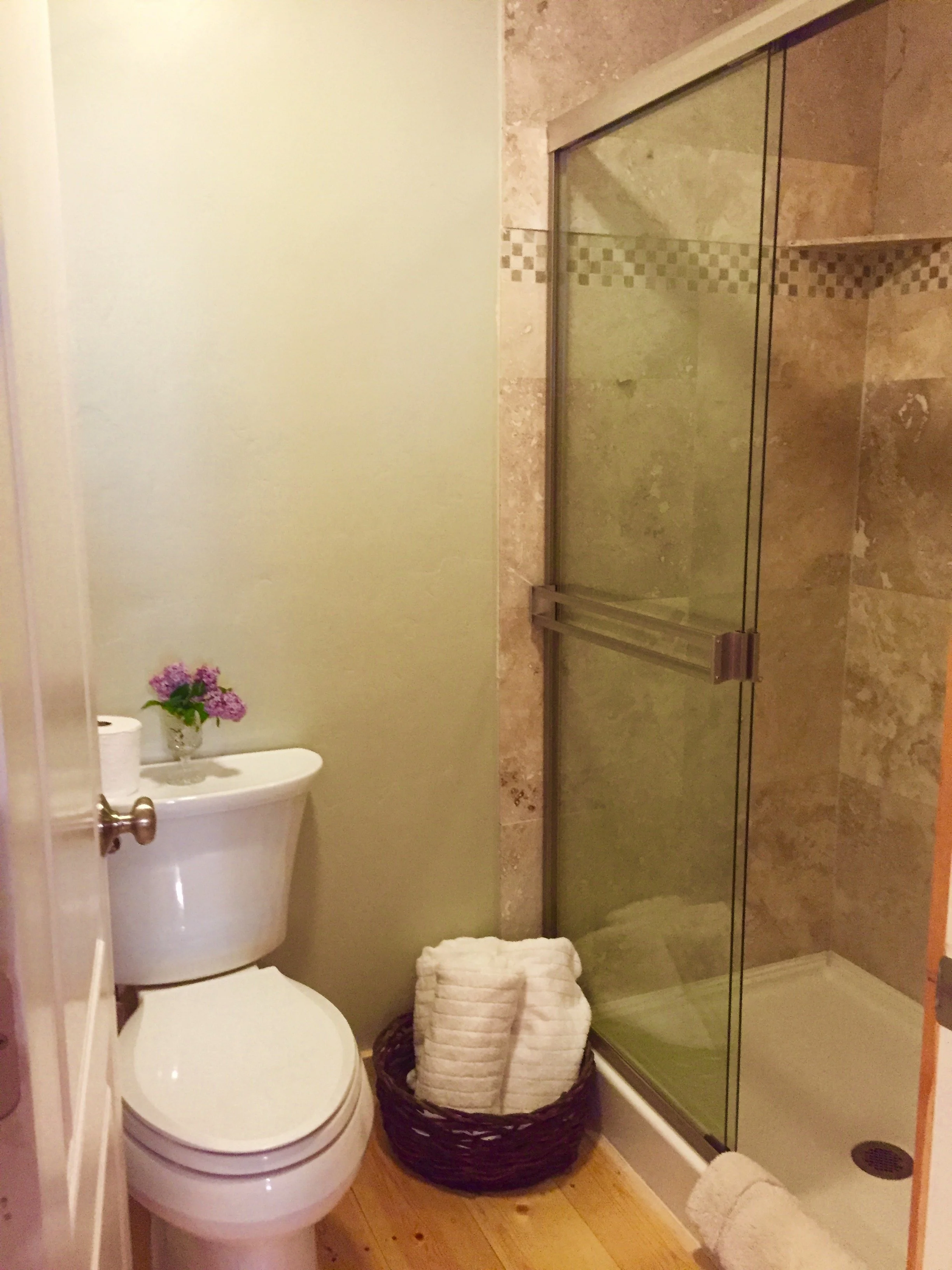 Small bathroom with a toilet, a basket of towels, and a shower with glass doors, beige tiles, and wooden floor.