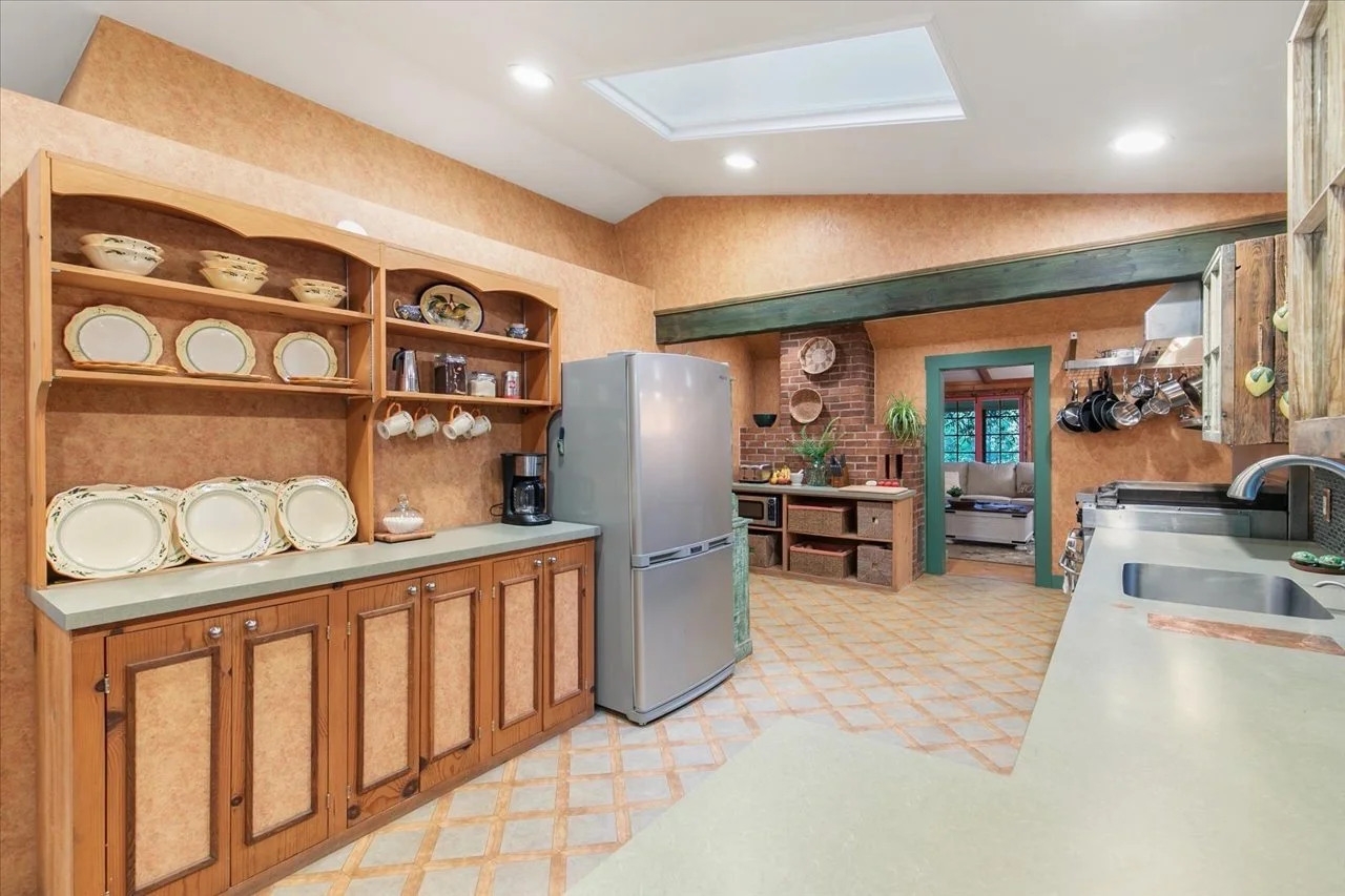 A kitchen with wooden cabinets, a stainless steel refrigerator, open shelves with dishes and mugs, a brick chimney, a countertop with a sink, and a view into a living room with curtains and a sofa.