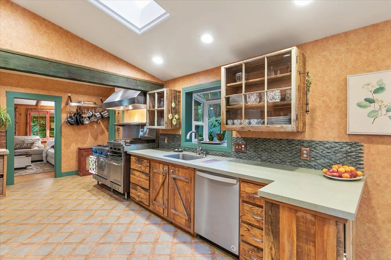 A rustic kitchen with wooden cabinets, a stainless steel stove, open shelving, a window above the sink, and beige tiled flooring. There is a doorway to a living room, and a bowl of peaches on the counter.
