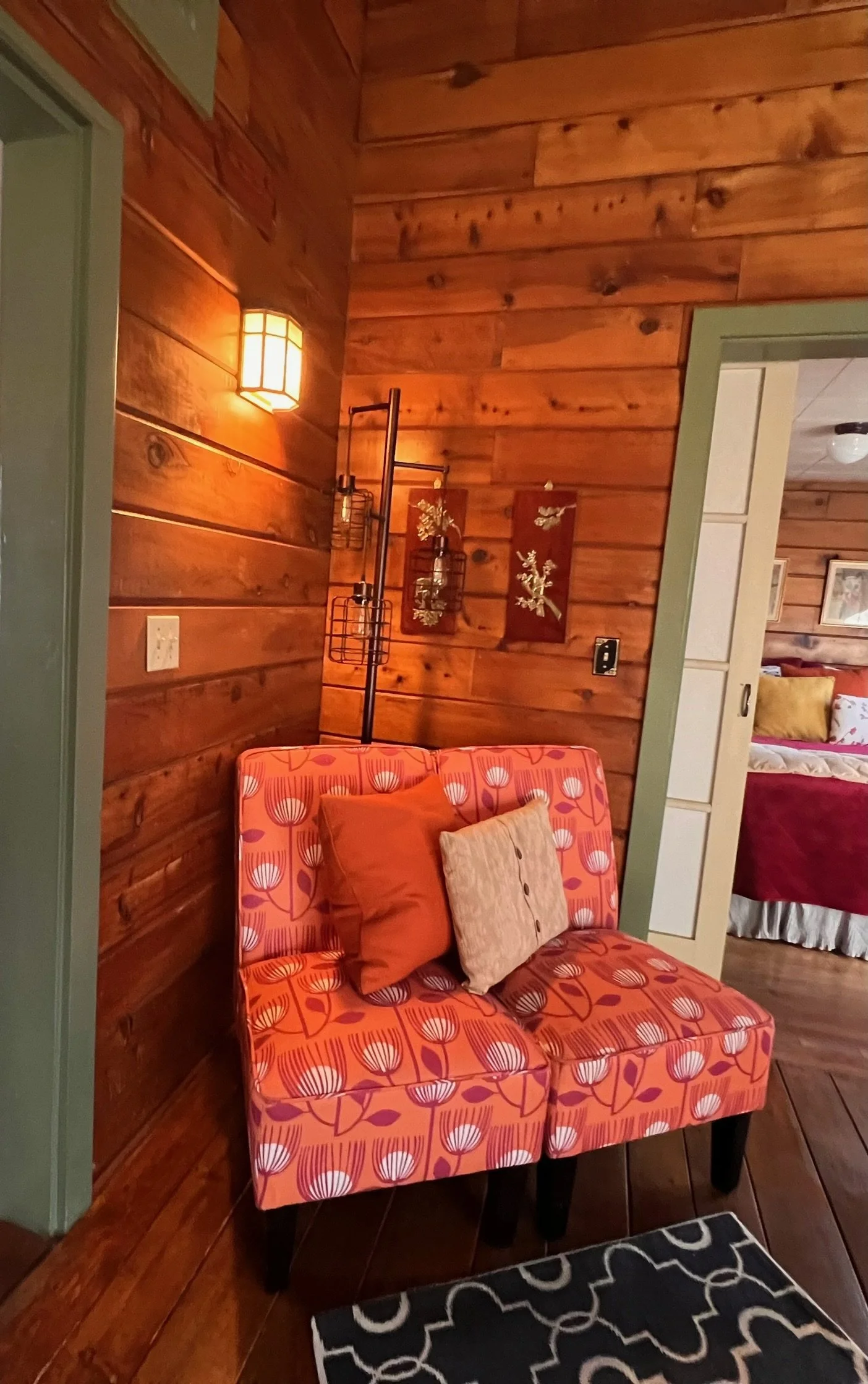 A cozy corner with a floral patterned orange loveseat, red and beige throw pillows, wood-paneled walls, warm lighting, and a doorway leading to a bedroom with a bed and framed artwork.