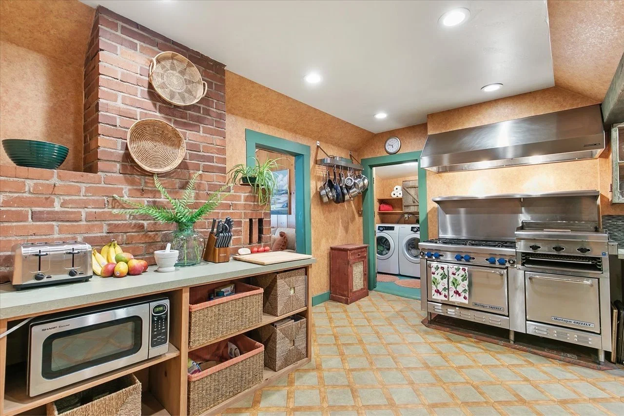 Kitchen with brick wall, wooden shelves, stainless steel appliances, laundry area with washer and dryer, and decorative woven baskets on the brick wall.