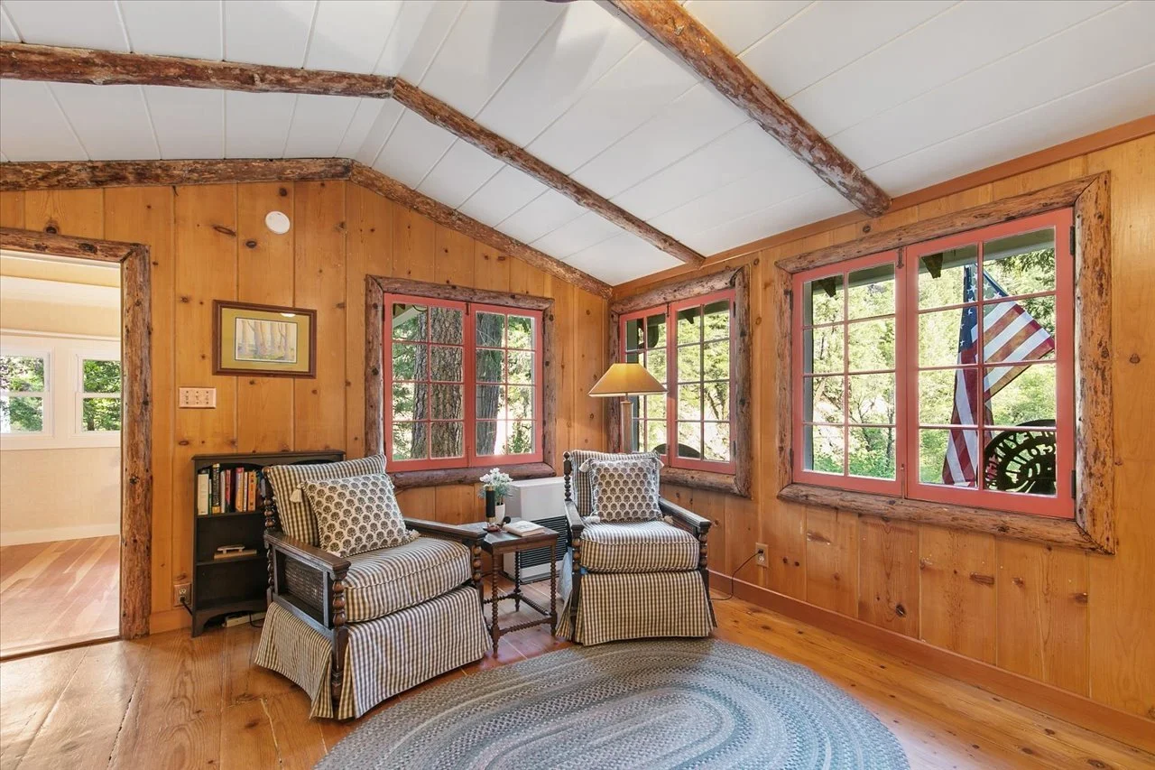 A cozy living room with wooden walls and ceiling beams, featuring two checked armchairs, a small table with a lamp and flowers, a bookshelf, and three large windows showing trees outside, with a view of an American flag outside.