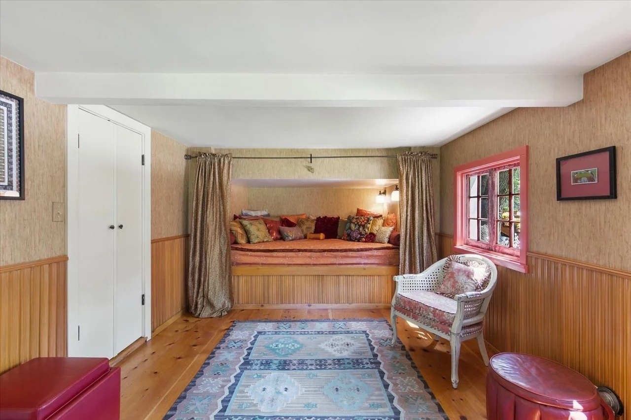 Cozy living room with wooden floors, beige wallpaper, and a built-in bed nook with colorful pillows and curtains. A window with pink trim lets in natural light, and wicker chair with a floral cushion is nearby. A rug covers part of the floor.