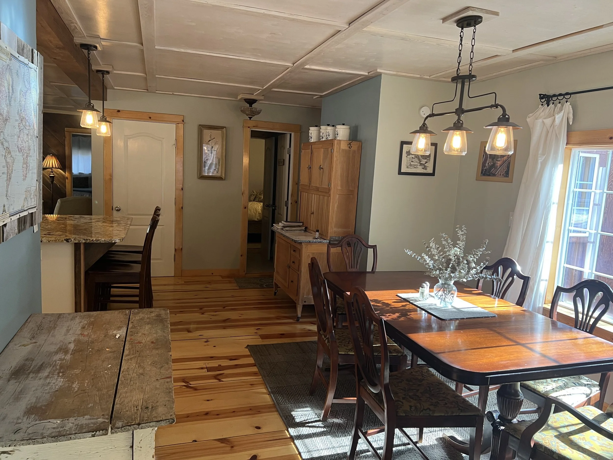 A cozy dining room with a wooden table and six chairs, a flower vase in the center, and a large window with white curtains letting in natural light. There is a wooden sideboard and a hanging light fixture with multiple bulbs above the table. The room
