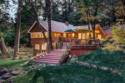 A large wooden house with a deck, surrounded by trees, with stairs leading down to a landscaped yard with rocks and grass.