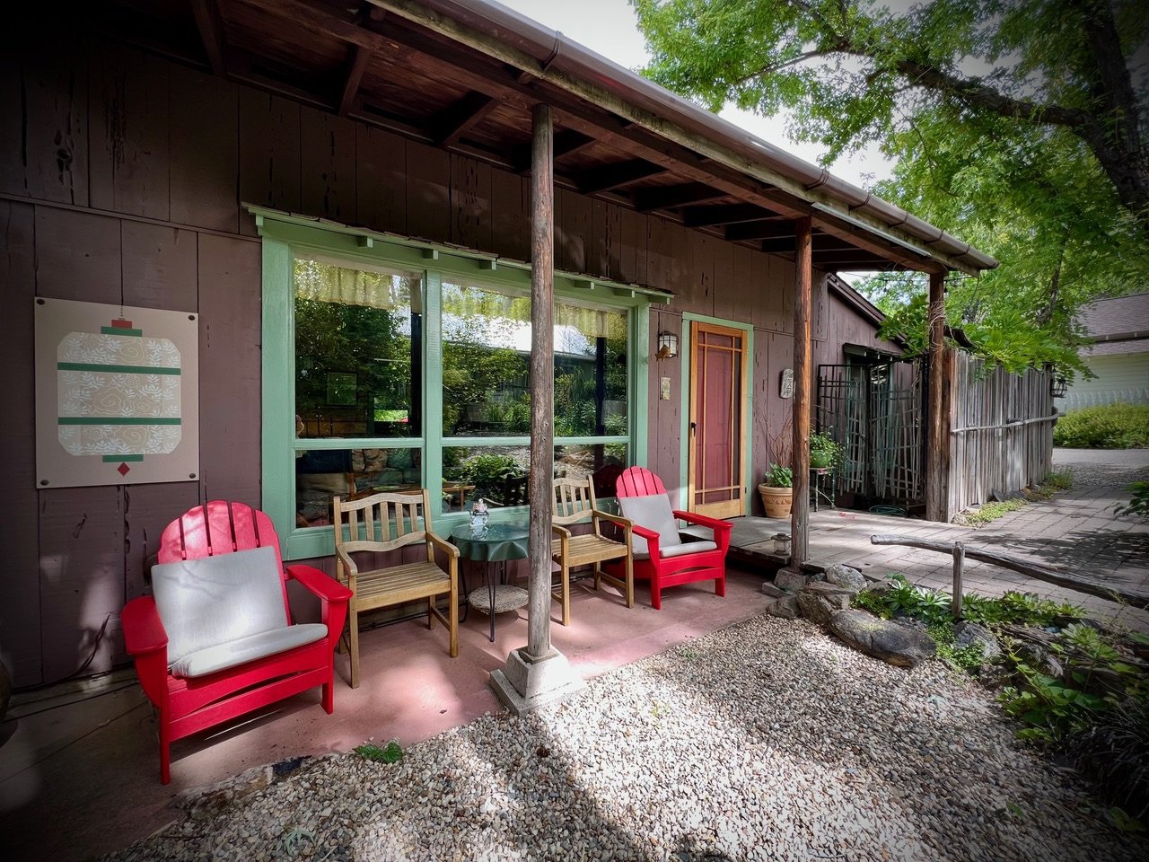 Front porch of a house with three chairs, one red and two wooden, around a small table, on a mix of pink concrete and gravel, with trees and fence in the background.