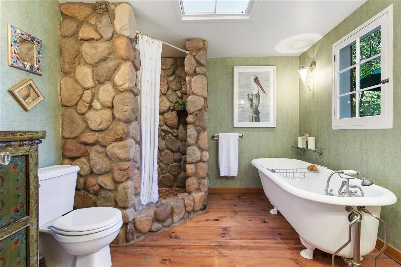 A rustic bathroom featuring a white clawfoot bathtub, a stone shower enclosure with a curtain, a toilet, and wooden flooring. The room has green walls, a skylight, and a window overlooking greenery. Decor includes framed artwork and small shelves wit