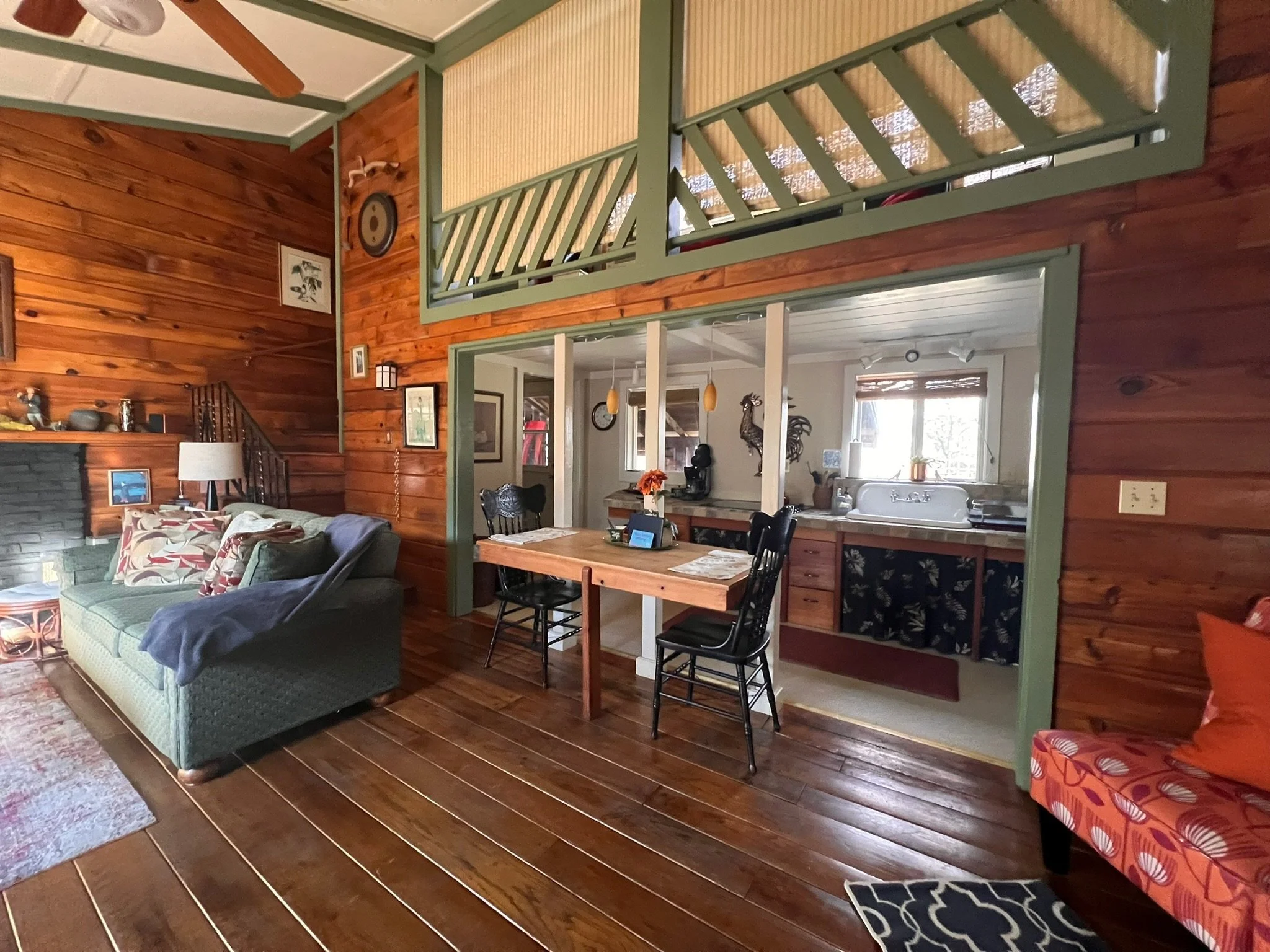 Interior view of a cozy living room with wooden walls and floors, featuring a green sofa, a patterned chair, a small table, and a view into a kitchen with a sink, window, and decor.