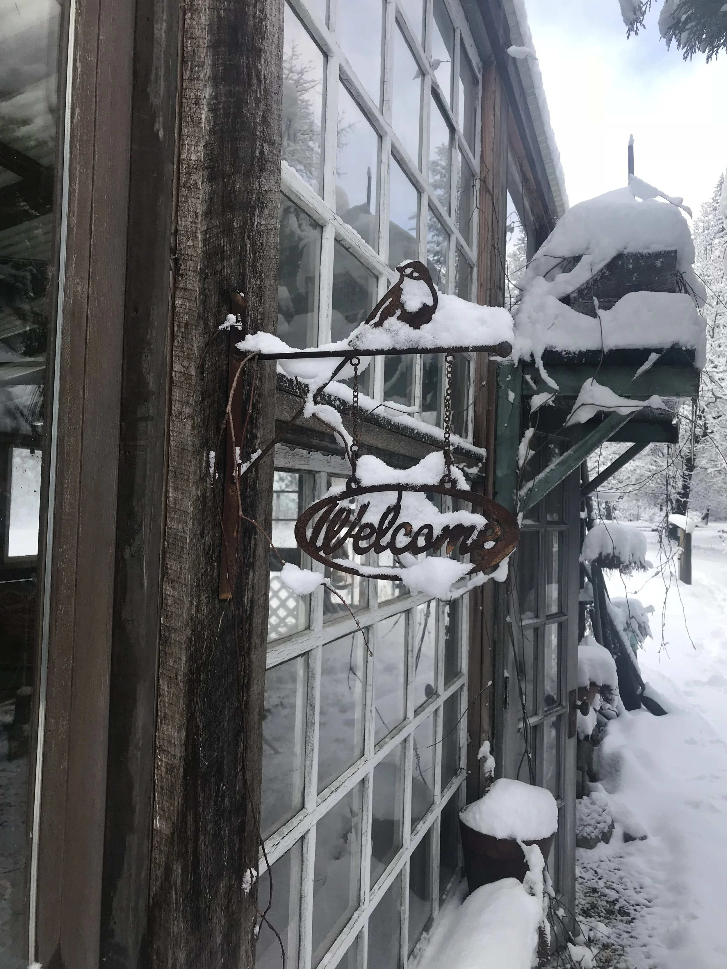 A snow-covered decorative welcome sign hanging on a rustic wooden building with large window panes. Snow accumulates on the sign, window ledge, and surrounding objects, indicating a snowy outdoor winter scene.
