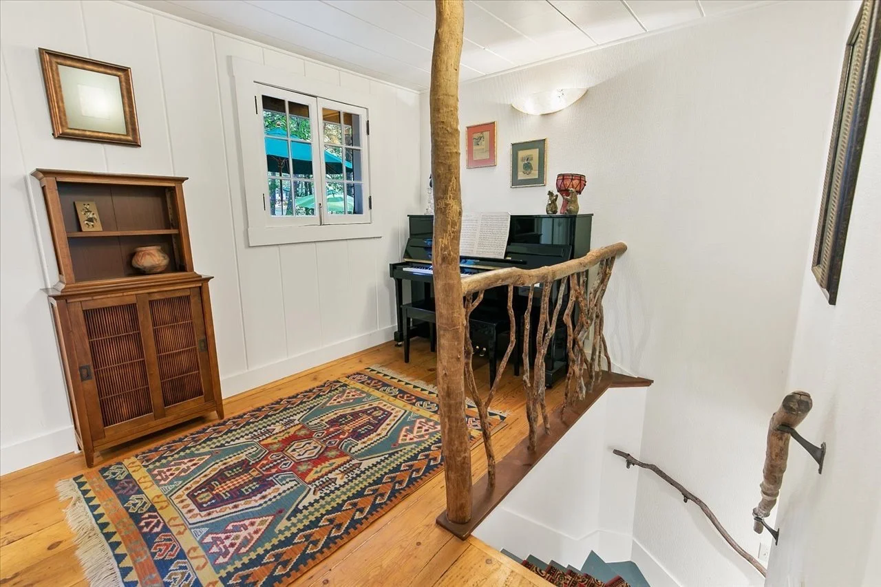 Interior view of a small upper landing area with a black upright piano, colorful framed artwork on the wall, and a decorative wooden railing made from tree branches. There is a window showing an outdoor patio with umbrellas, a wooden cabinet, and a p