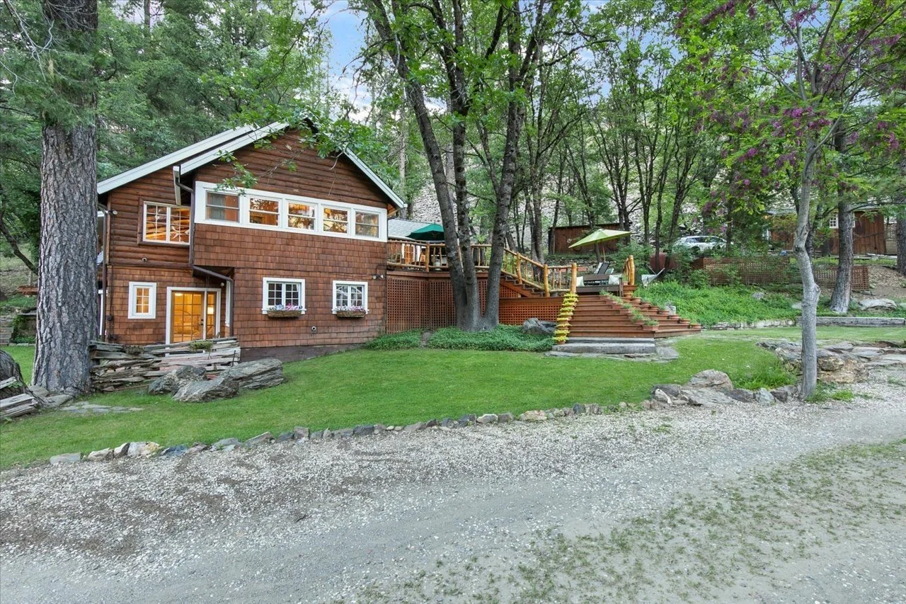 A wooden house on a grassy yard, surrounded by trees, with a deck and outdoor dining area.