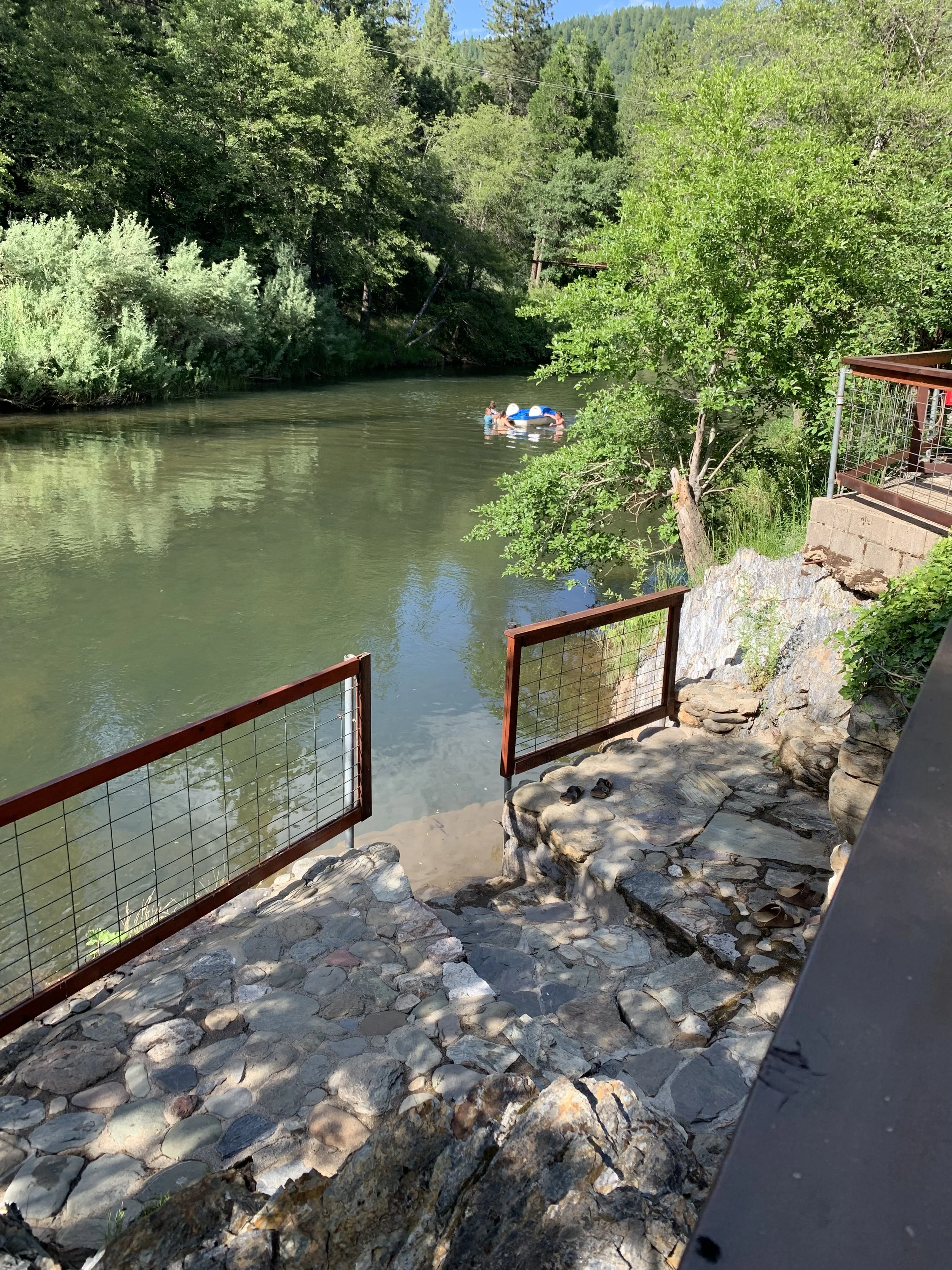 View of a river with green trees on both sides, a small boat with people on it, and a rocky stairway with metal railing in the foreground.