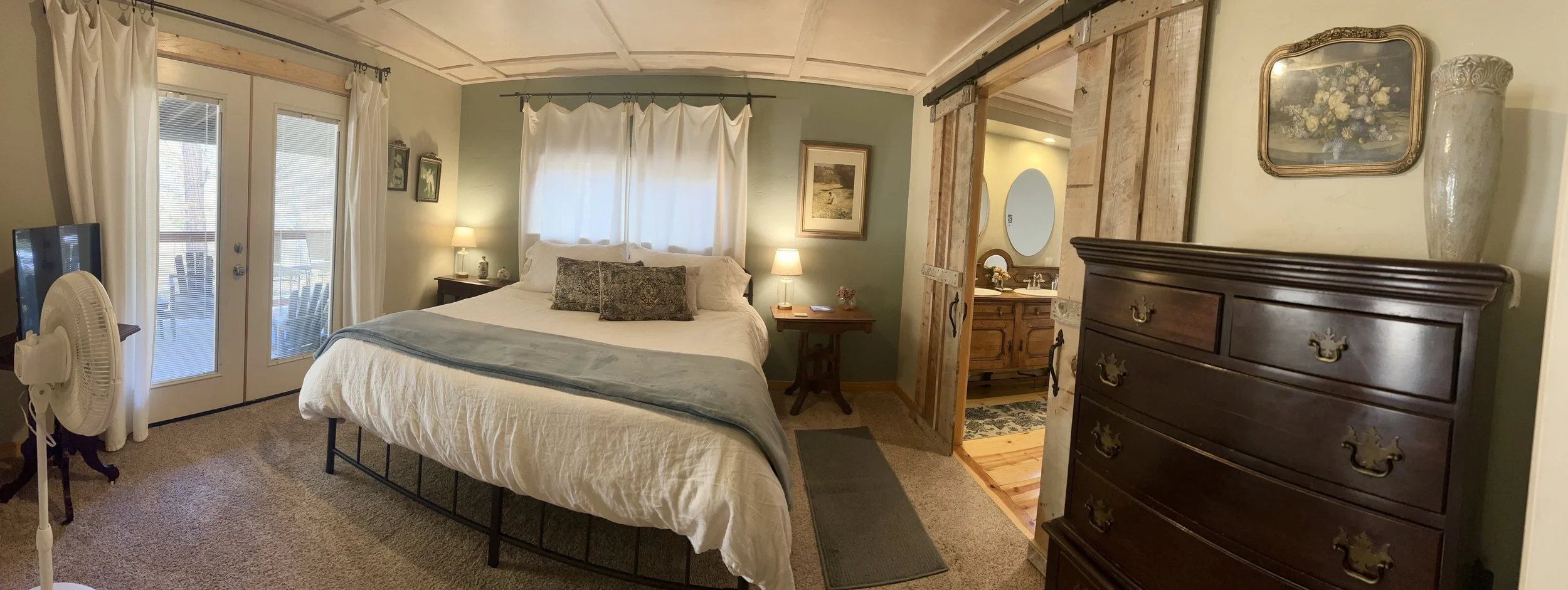 A cozy bedroom with a large bed covered in white and light blue bedding, flanked by nightstands with lamps, a dresser on the right, and a sliding barn door leading to a bathroom. There are two glass doors with white curtains leading to an outdoor dec