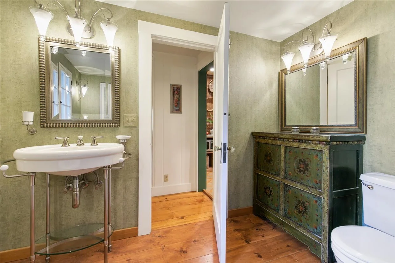 A vintage bathroom with green textured walls, wooden flooring, a white porcelain sink with metal legs, a large mirror with a striped frame, and a green wooden cabinet with floral patterns and a mirror above.