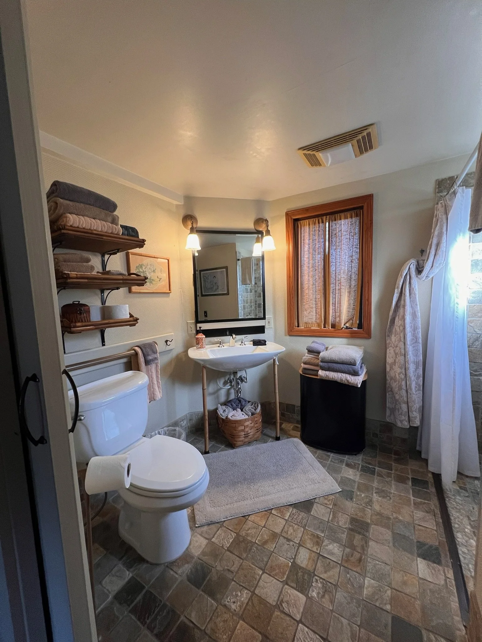 Bathroom with a toilet, small sink, mirror, window with curtains, and shelves with towels.