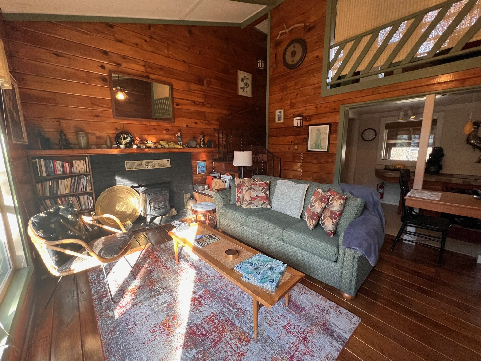Living room with wooden walls, a green sofa with patterned cushions, a wooden coffee table with books, an armchair, a painted rug, a brick fireplace with decorative items, and various pictures on the wall.