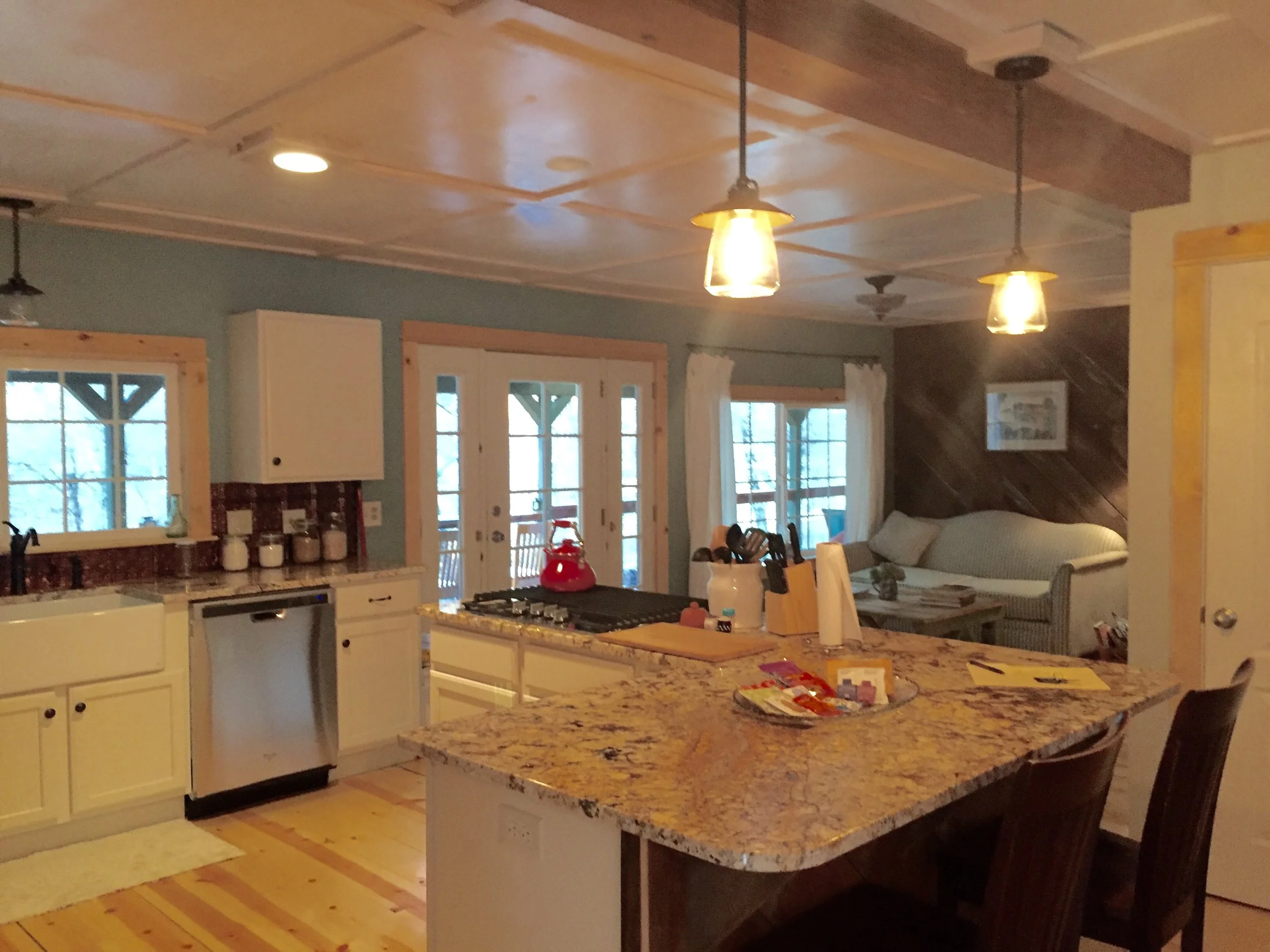 A cozy kitchen and living area with a granite countertop island, white cabinets, and a stove with a red kettle. There is a doorway with glass doors leading outside, and a living space with a couch and framed artwork on a wooden accent wall.