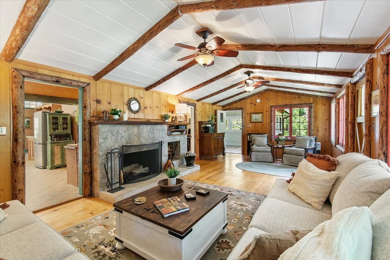Living room with wood-paneled walls, exposed wooden beams, a stone fireplace, ceiling fans, a beige sofa, armchairs, windows with view outside, and a wooden coffee table.