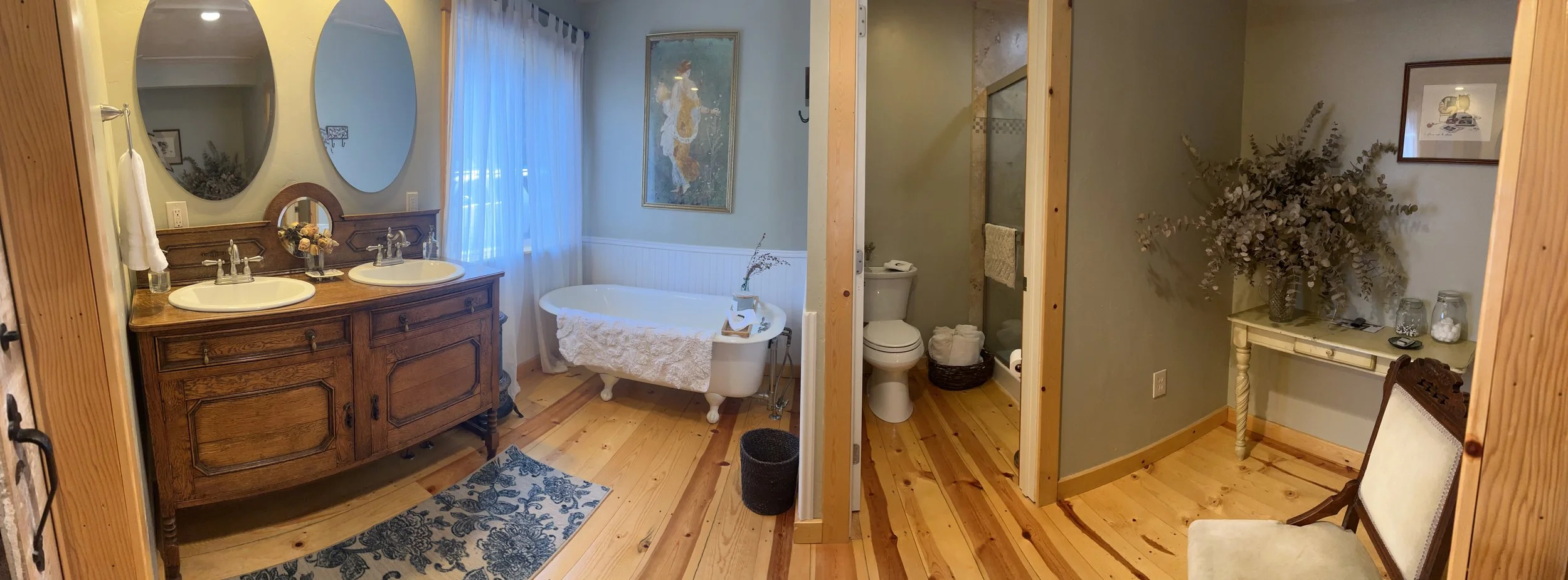 A bathroom with a double sink vanity, a bathtub, a toilet, and a small table with a large vase of dried flowers. The bathroom has wooden floors and light blue walls with framed art and curtains.