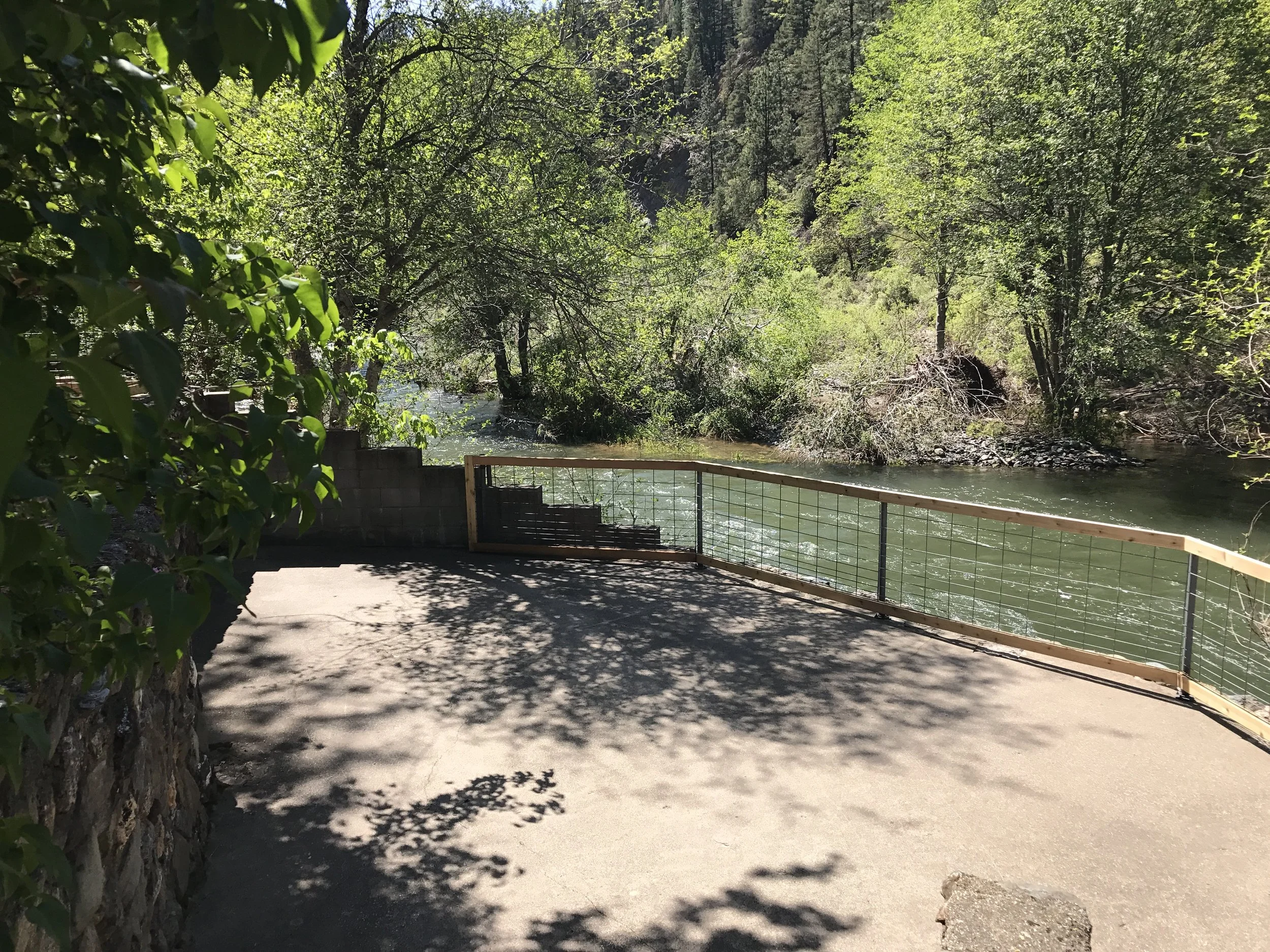 A riverside viewing platform with a metal railing surrounded by trees and greenery, overlooking a flowing river.