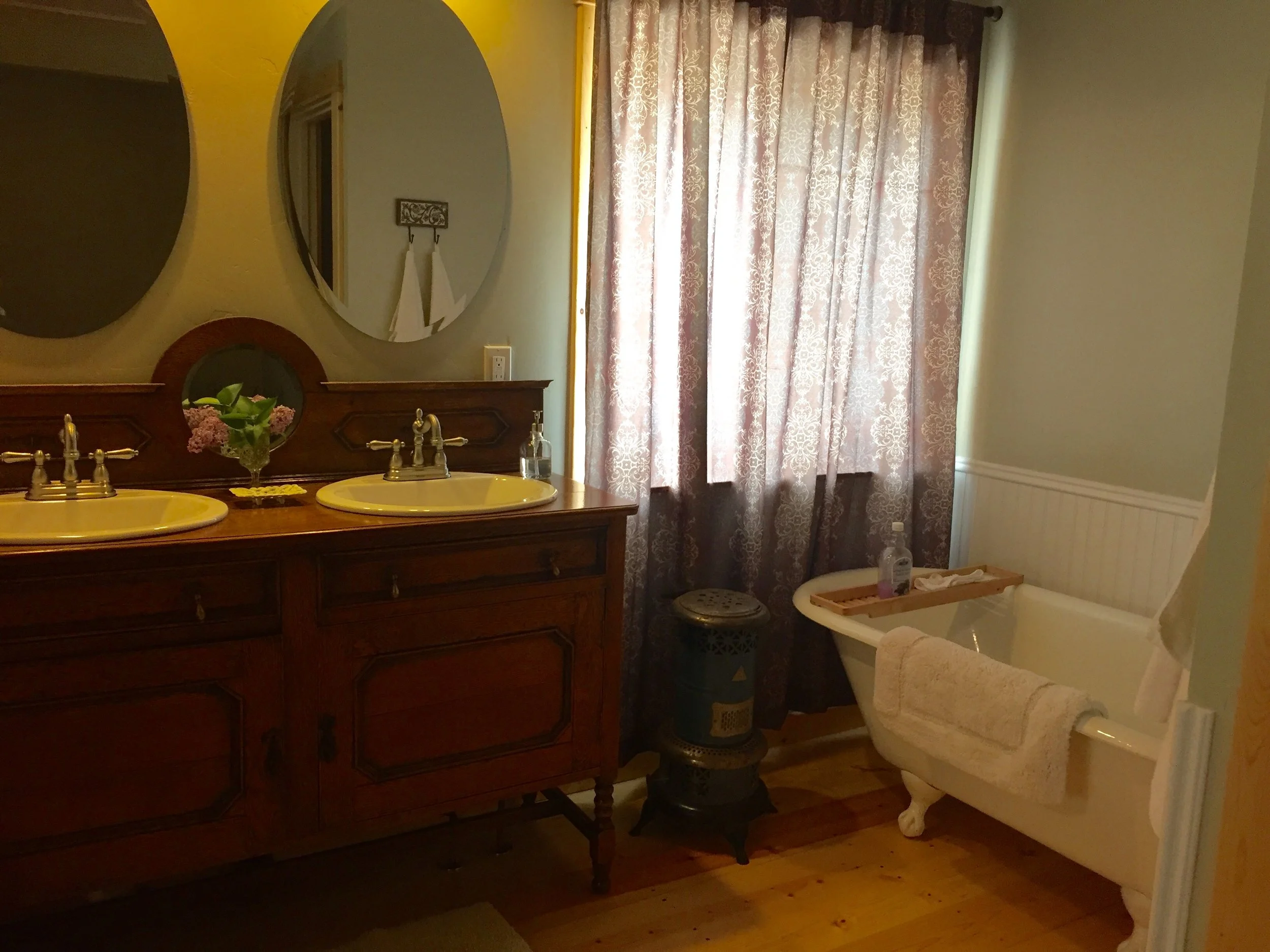 A bathroom featuring a vintage wooden double sink vanity with two mirrors above, floral arrangements, and a soap dispenser. There is a clawfoot bathtub with a towel hanging over its edge, a small side table, and a window with patterned curtains, all 