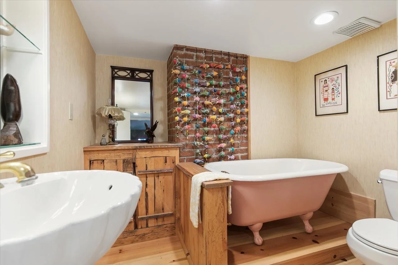 A bathroom with a pink clawfoot bathtub, a wooden vanity, a white sink, and a brick wall decorated with hanging colorful balls and fabric, along with framed artwork on the beige walls.