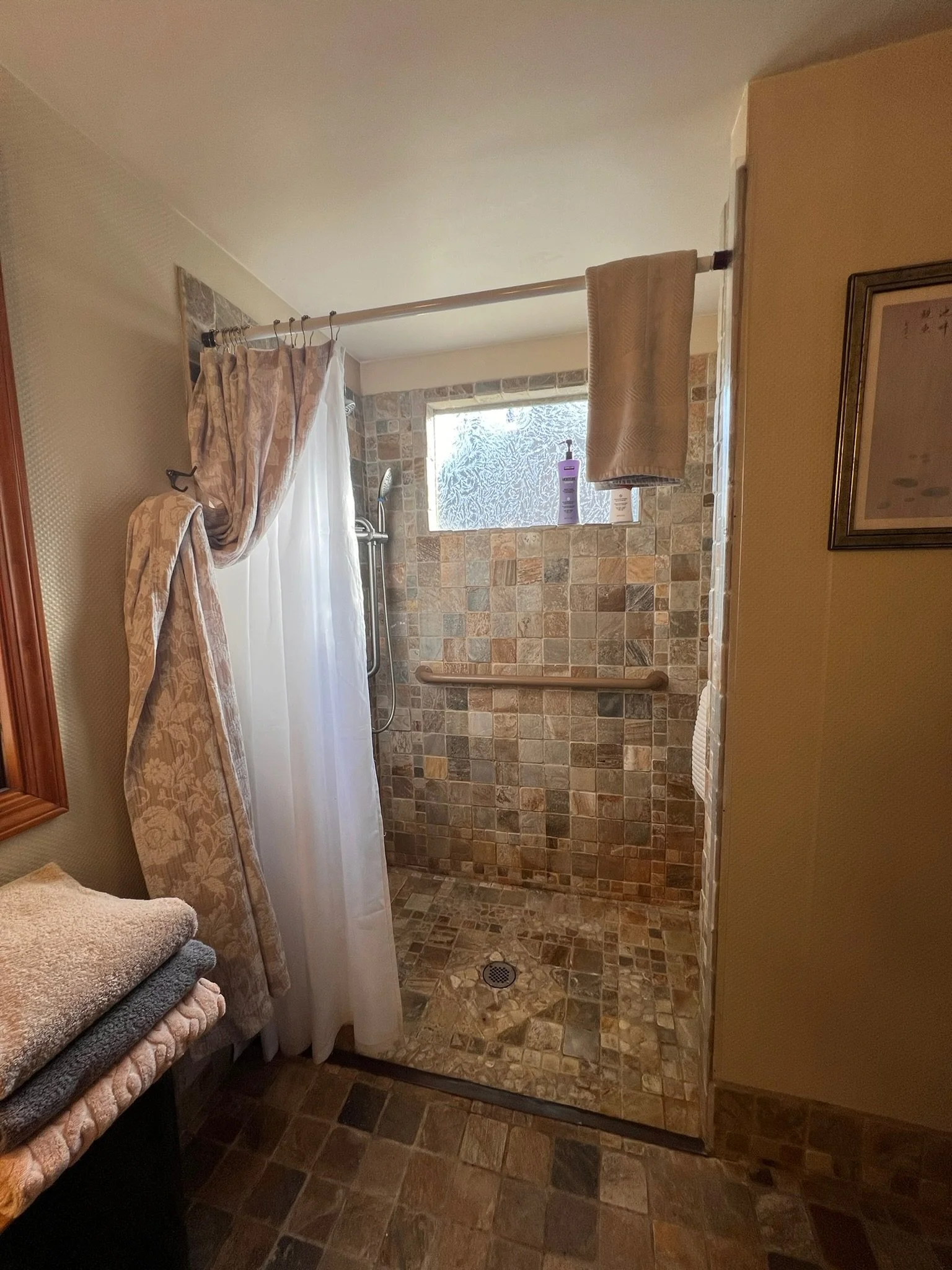 Bathroom shower area with brown and beige tile walls, frosted window, and a grab bar. Shower curtain partially drawn, folded towels on a shelf, and a framed picture on the wall.