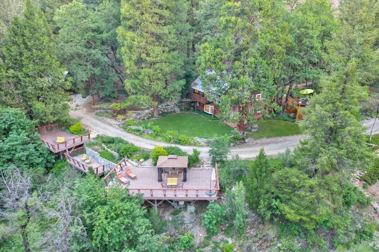 Aerial view of a house surrounded by tall trees, with a large deck featuring outdoor furniture and lounging areas. A grassy yard and a dirt road are visible around the property.
