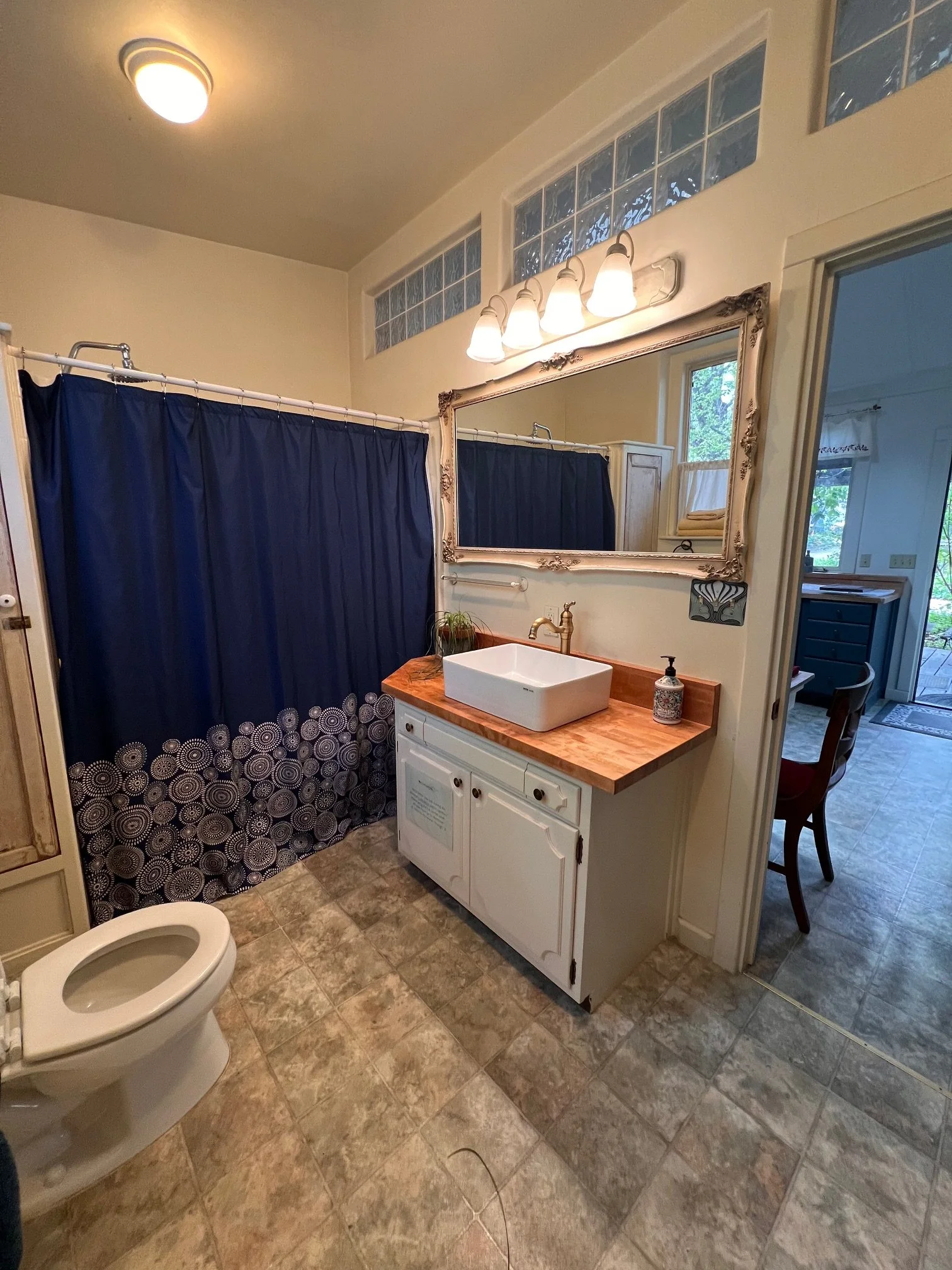 A bathroom with a toilet, a vanity with a sink, a large wall mirror, blue shower curtain, and a light fixture with four bulbs.