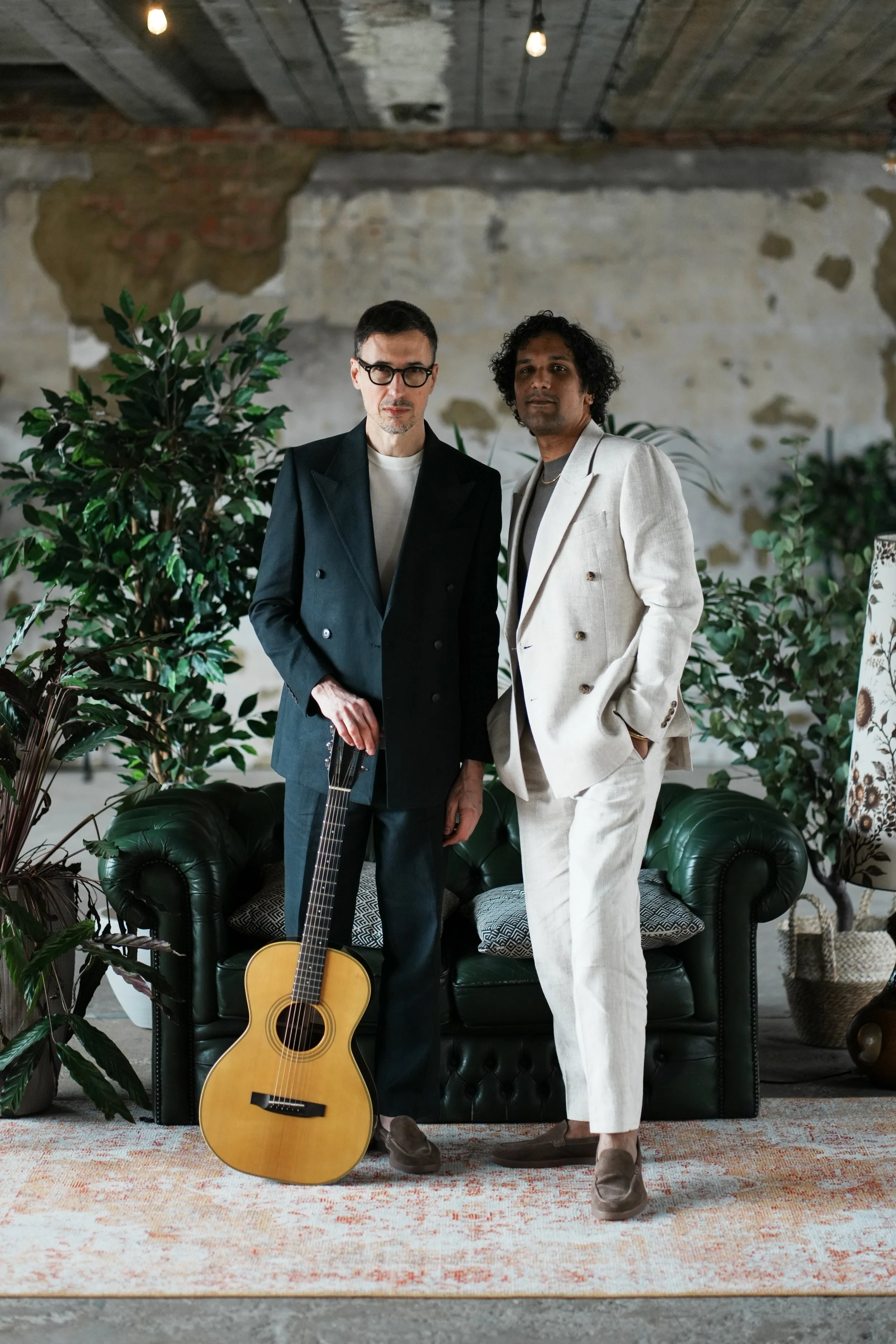 Two men in formal wear standing in front of a green leather sofa, surrounded by indoor plants. One holds a guitar. The setting appears to be an indoor space with a rustic, unfinished wall and ceiling, and soft lighting.