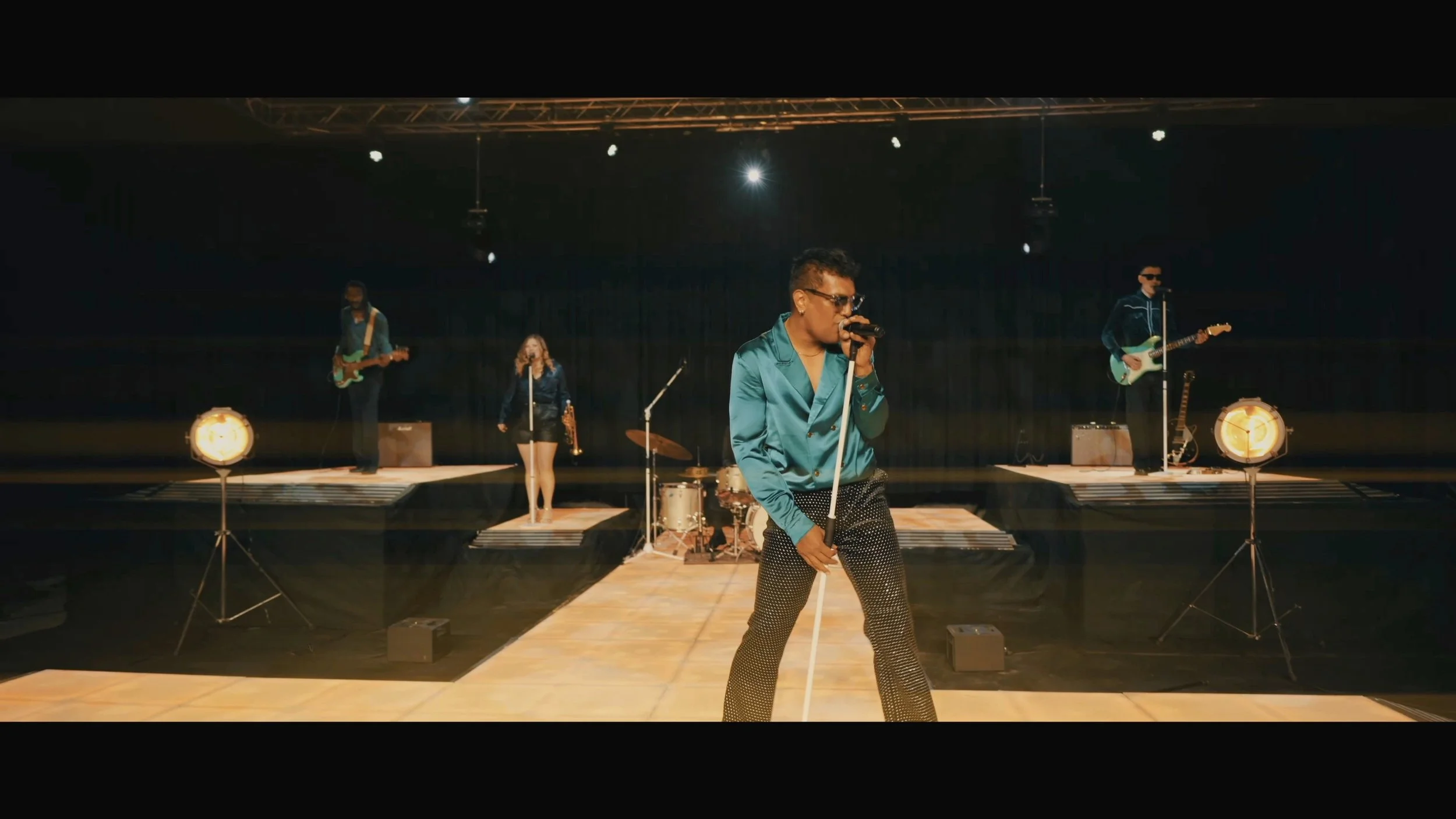 A band performing on a stage with a singer in front, holding a microphone, wearing sunglasses, a shiny teal shirt, and black pants with white dots. In the background, there are two guitarists, a female vocalist, and a drummer. The stage is lit with warm lighting.