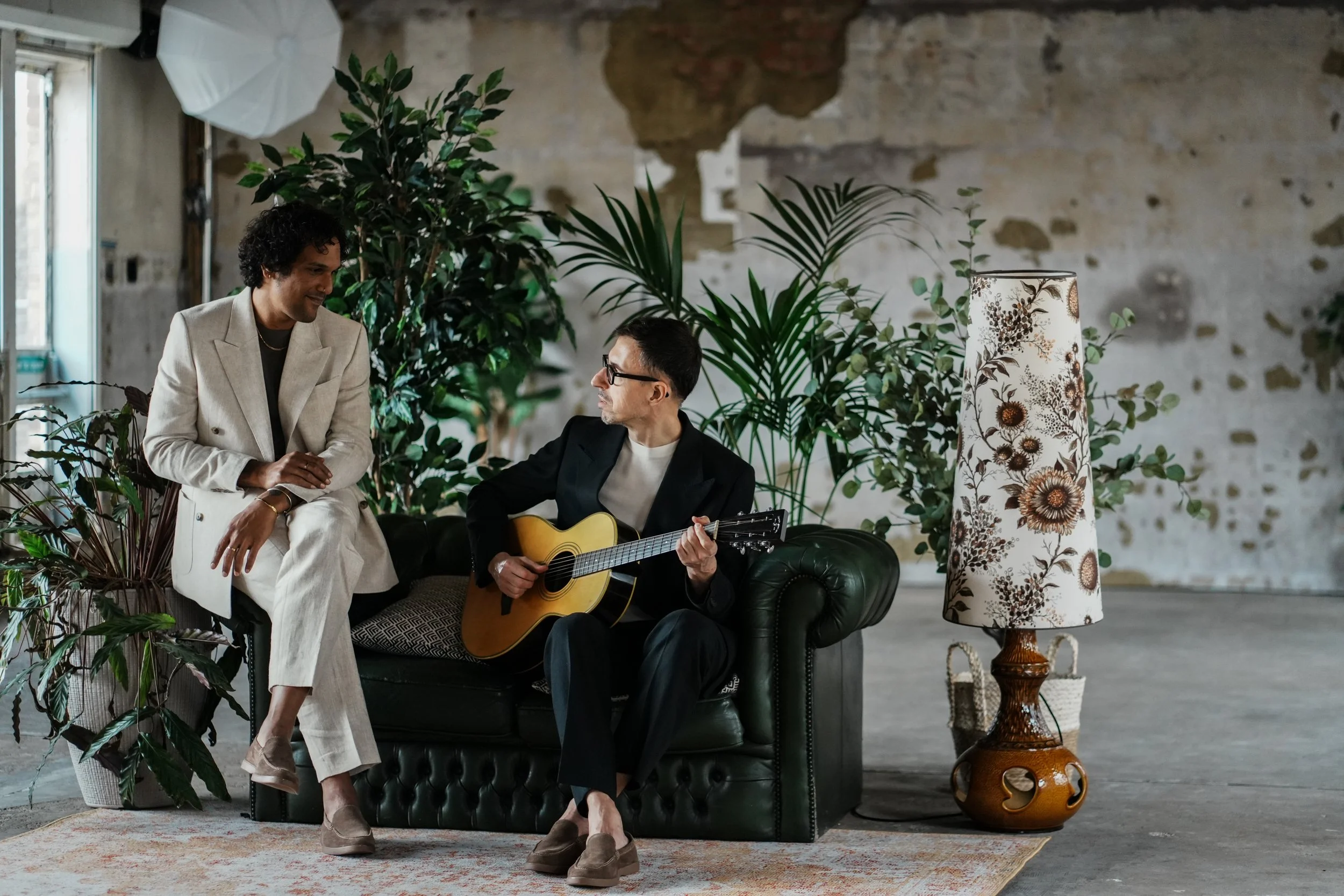 Two men in a cozy, industrial-style room with plants and a vintage lamp. One man in a light-colored suit sits on a black leather sofa, smiling and listening. The other man in a dark suit plays an acoustic guitar, sitting on the sofa.