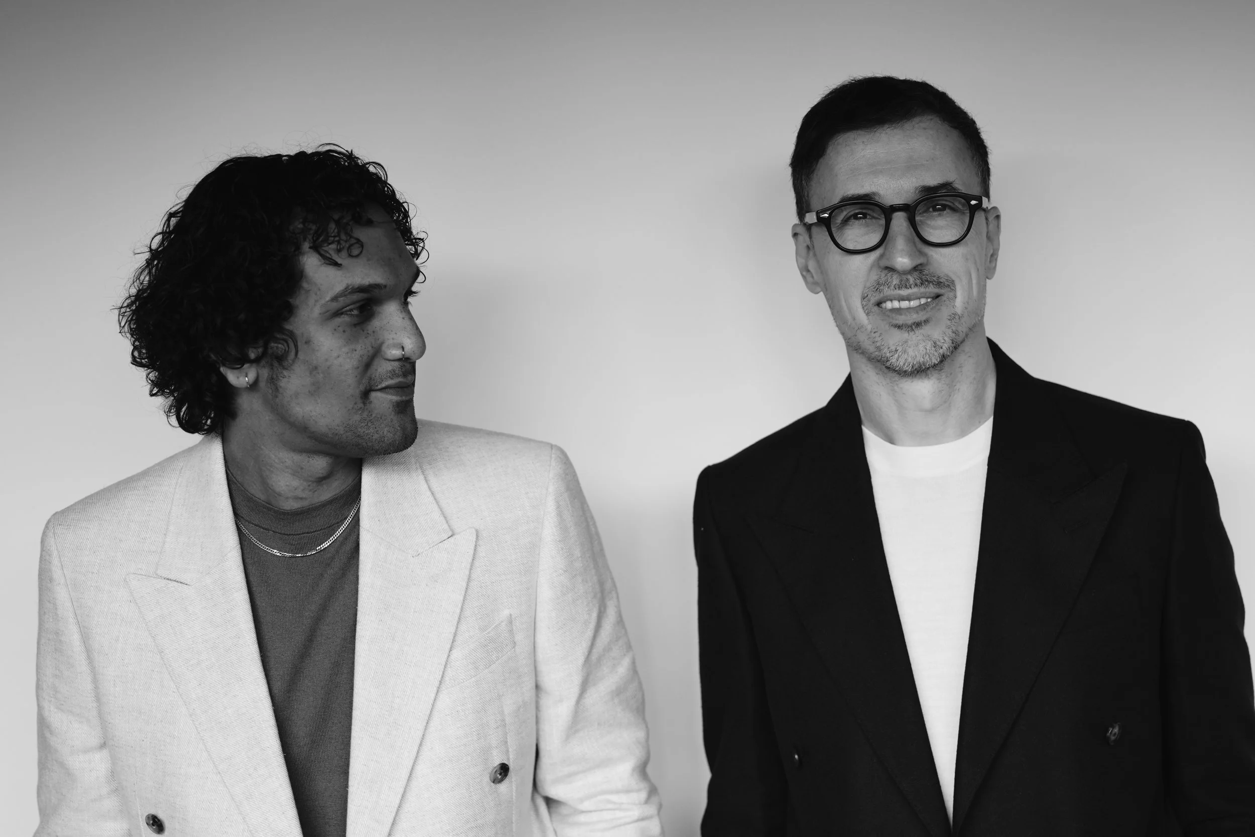 Two men standing side by side, one looking at the other and smiling, both dressed in suits, against a plain background.