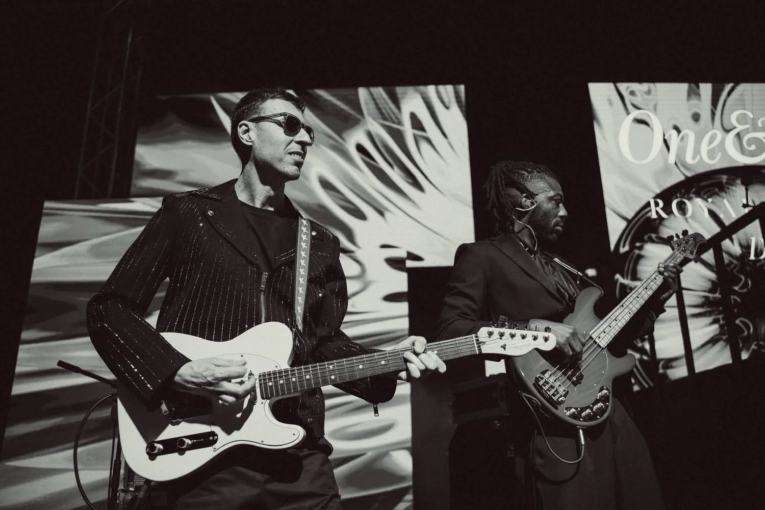 Two musicians playing electric guitars on stage with abstract backgrounds projection.