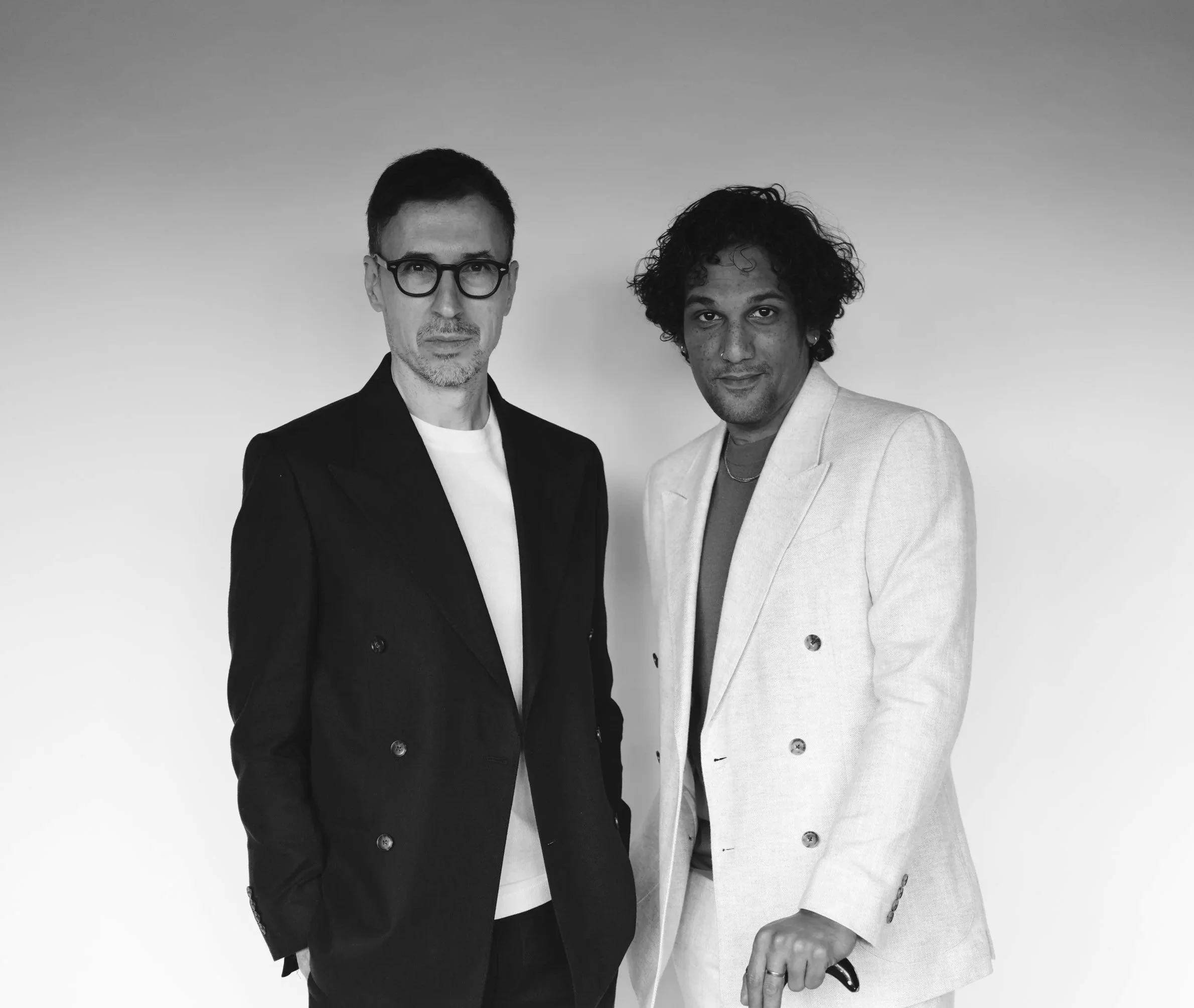 Two men standing side by side against a plain background, dressed in blazers and casual shirts.